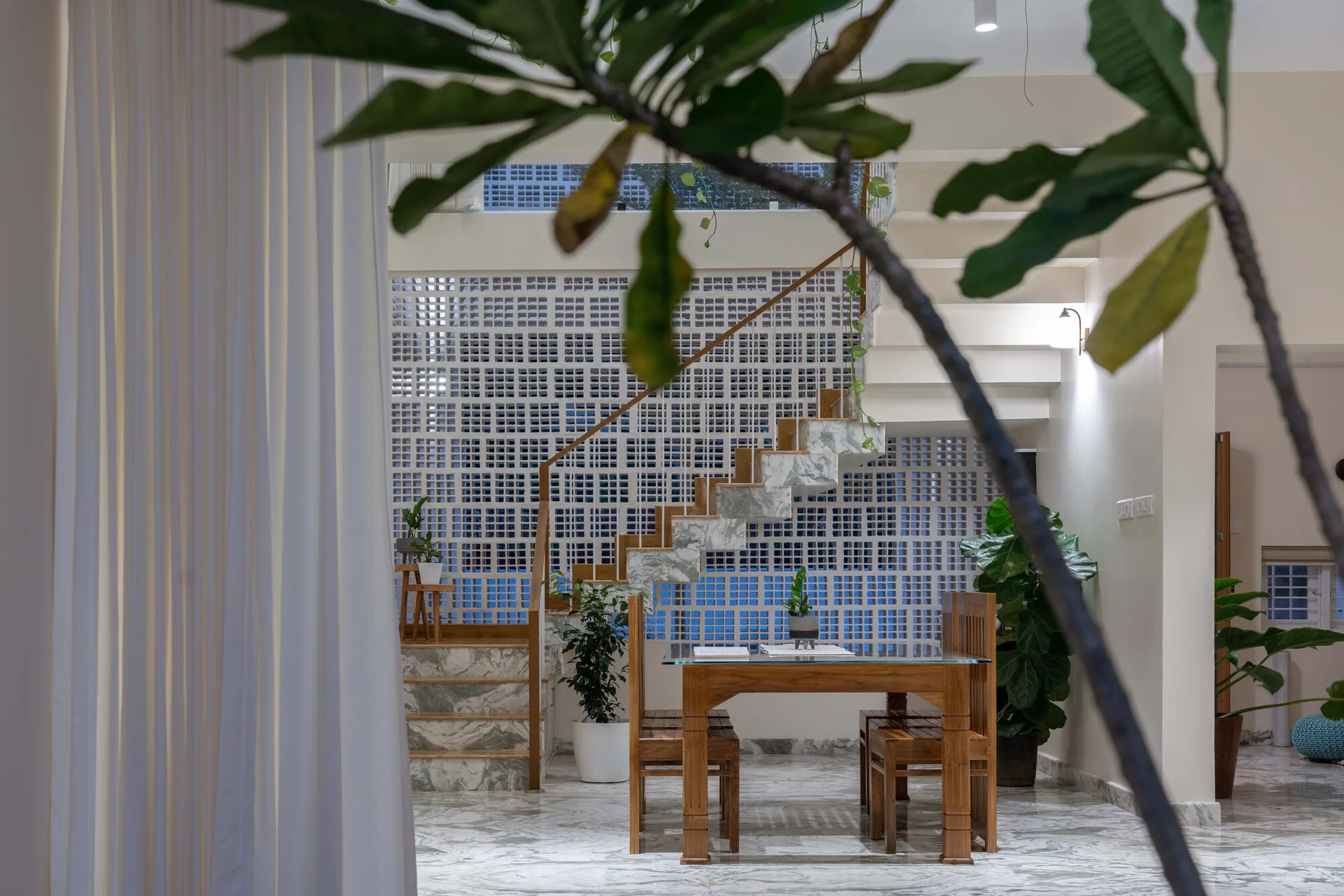 A wooden dining set stands before a floating marble staircase and a white breeze block screen wall in a bright, modern interior.