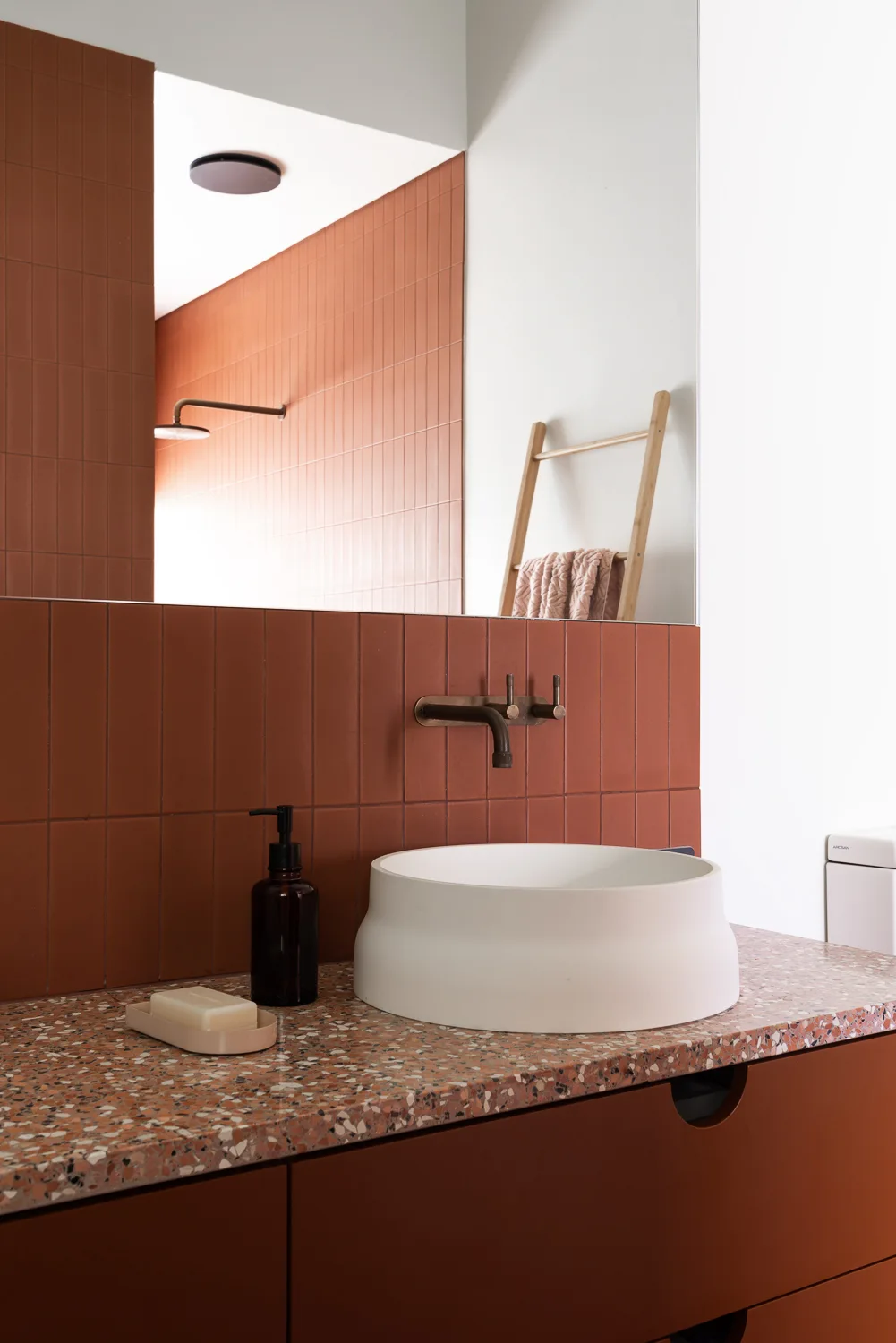 A white vessel sink sits on a terrazzo vanity countertop against a vertically tiled terracotta wall with a wall-mounted bronze faucet.