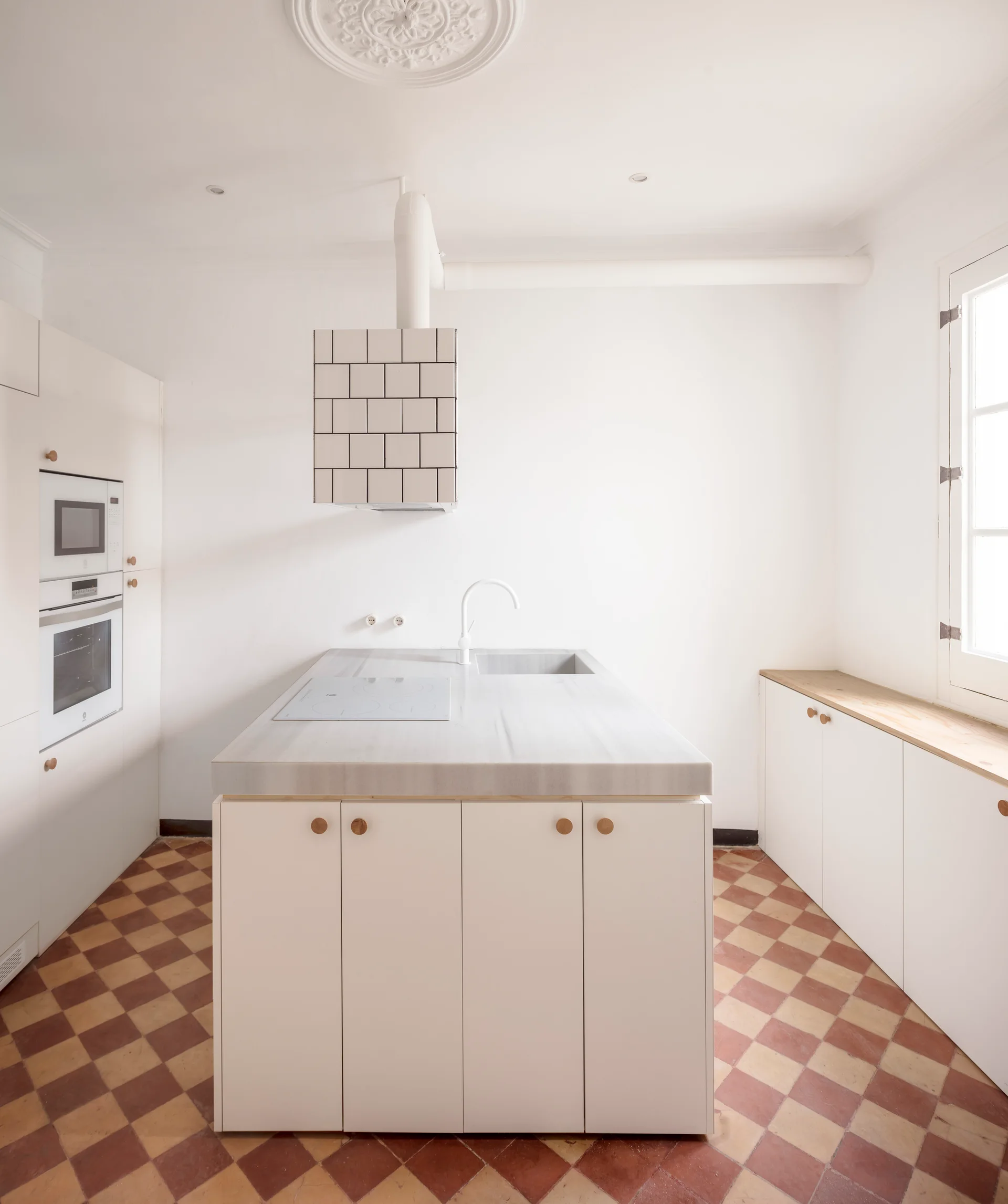 A bright minimalist kitchen featuring a stone-topped island, white cabinetry with wood knobs, and a terracotta checkerboard tiled floor.