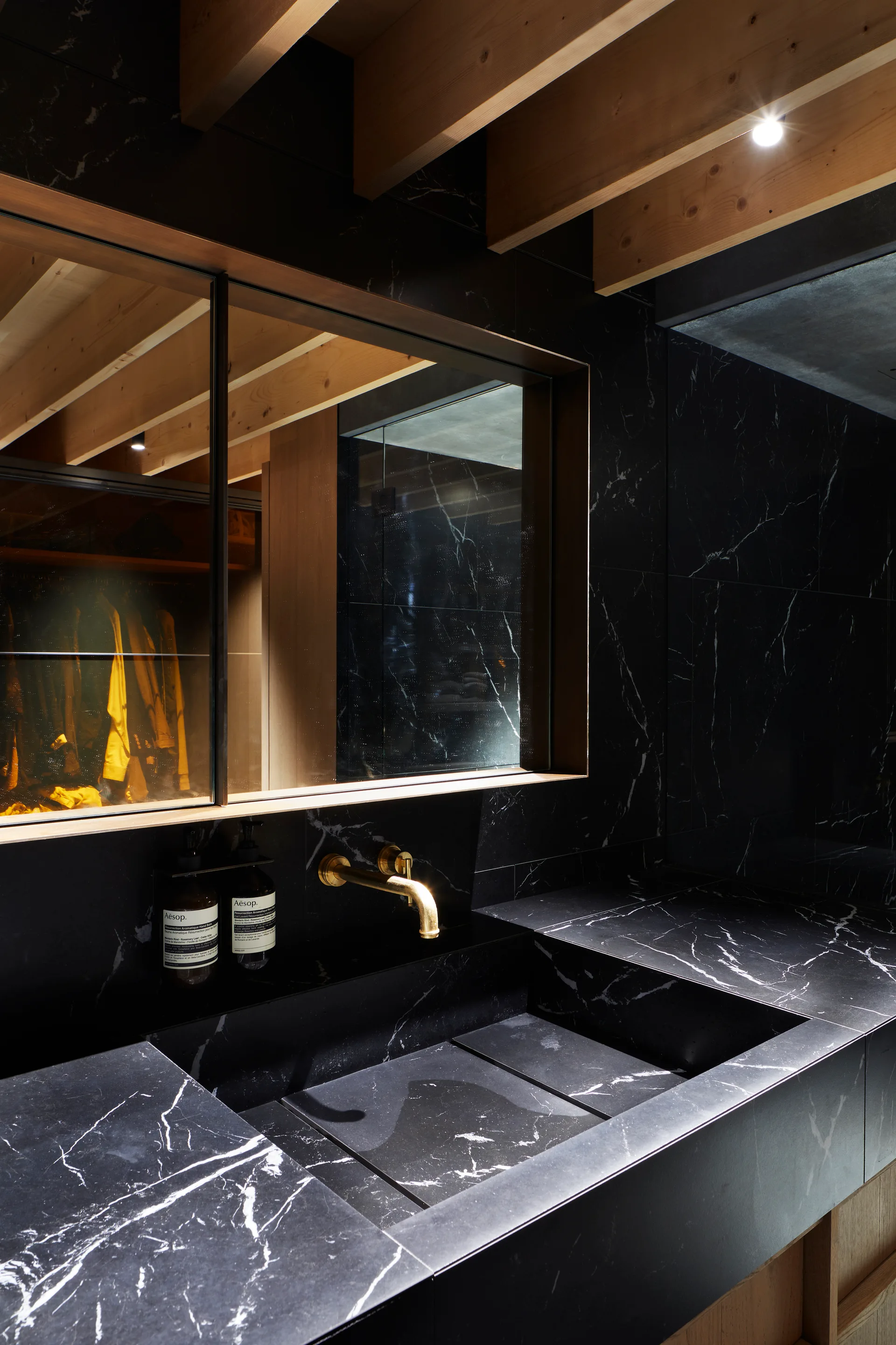 A modern bathroom vanity featuring an integrated black marble sink, wall-mounted brass faucet, and mirror beneath exposed wood ceiling beams.