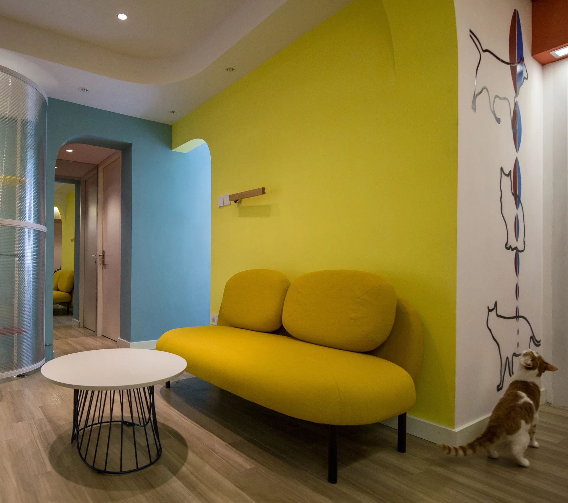 A modern yellow sofa sits against a matching yellow wall next to a blue wall with arched doorways and a round white coffee table.