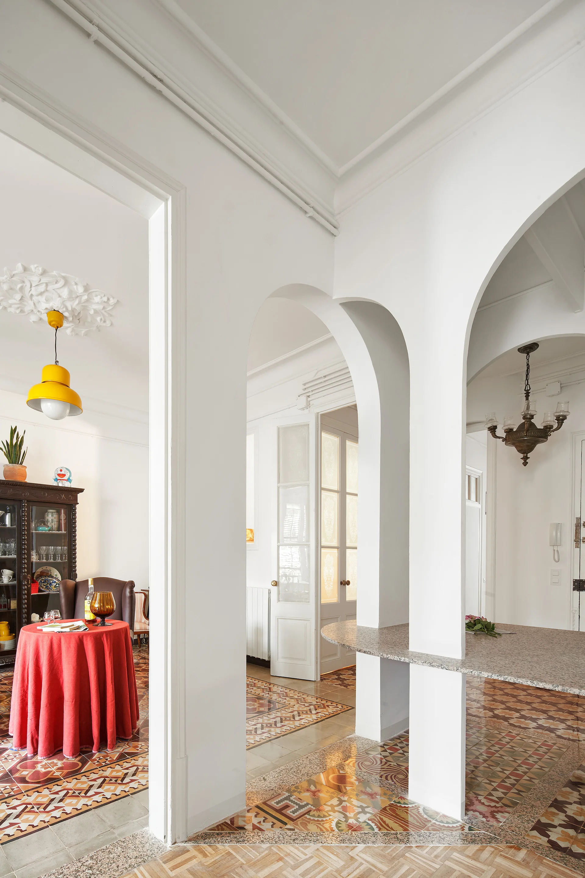 A bright white interior with large plaster archways, colorful geometric patterned floor tiles, and a round table dressed in a red tablecloth.