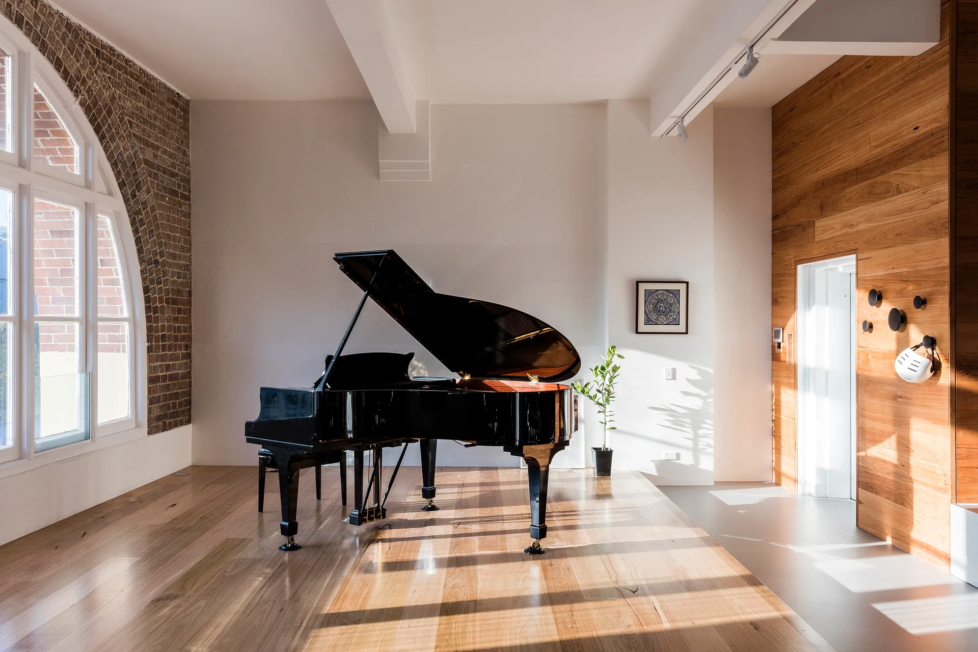 A black grand piano stands on light timber flooring next to a large arched window with exposed brick framing.