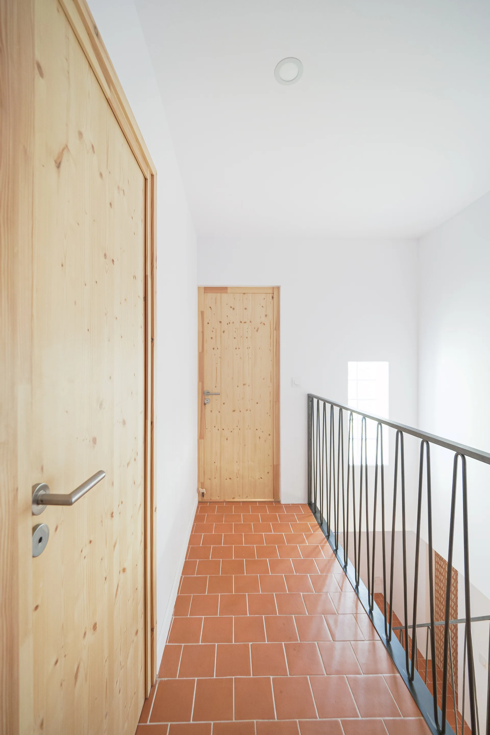 A bright hallway featuring rectangular terracotta floor tiles, white walls, natural knotty wood doors, and a slim black metal staircase railing.