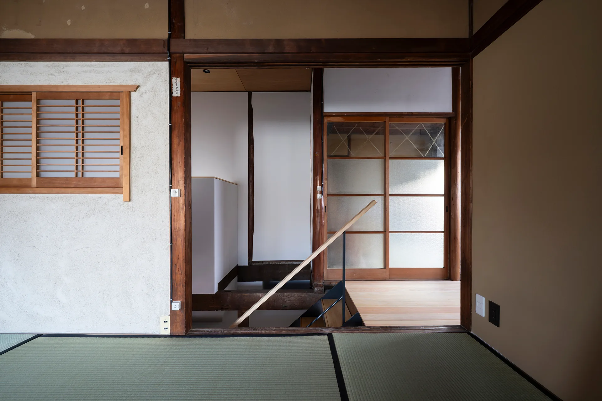 A modern staircase with a slim wooden handrail and black metal post descends through tatami flooring in a traditional Japanese room.