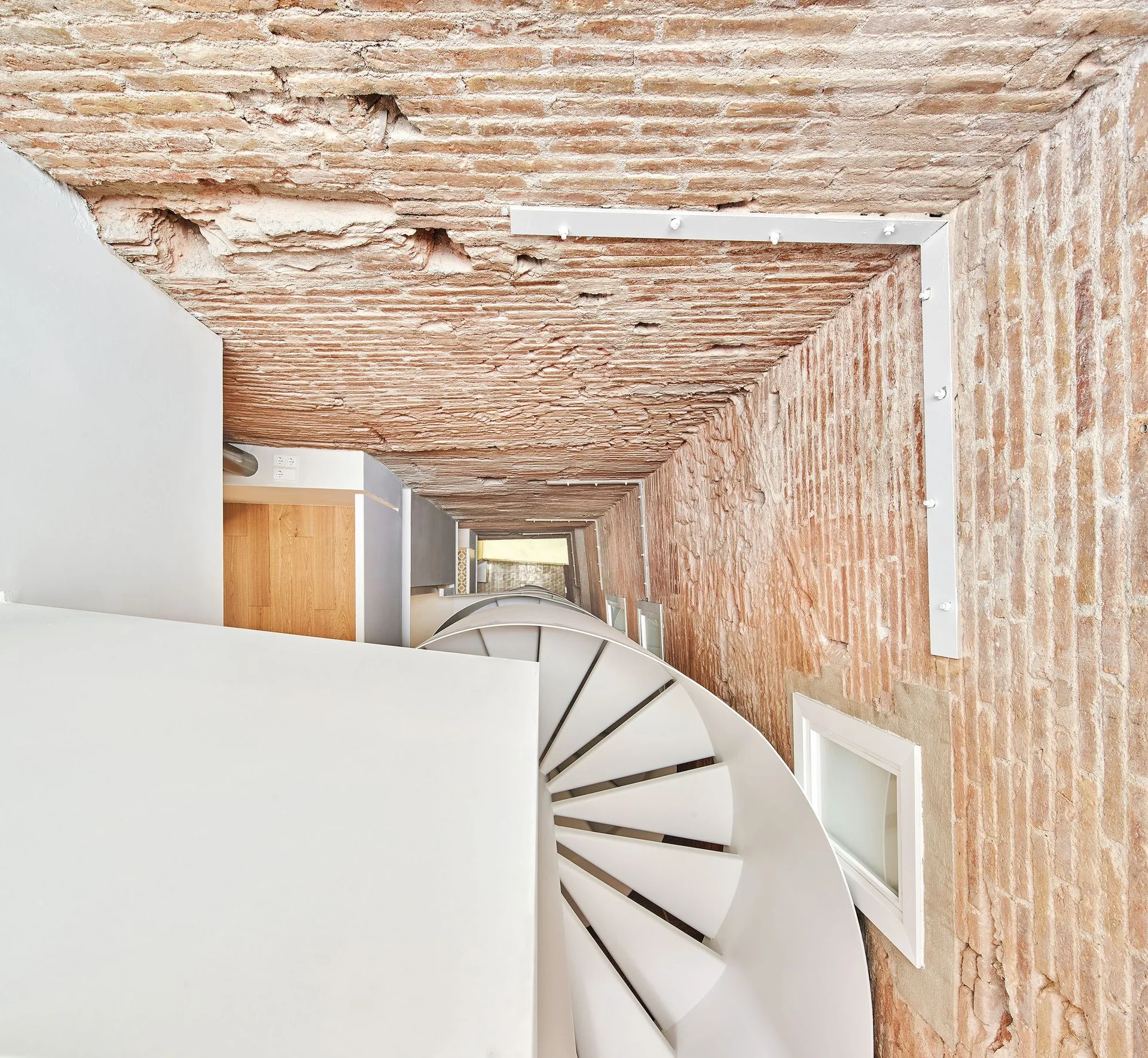 A high-angle view looking down a narrow stairwell with a white metal spiral staircase set against rustic exposed brick walls and ceiling.