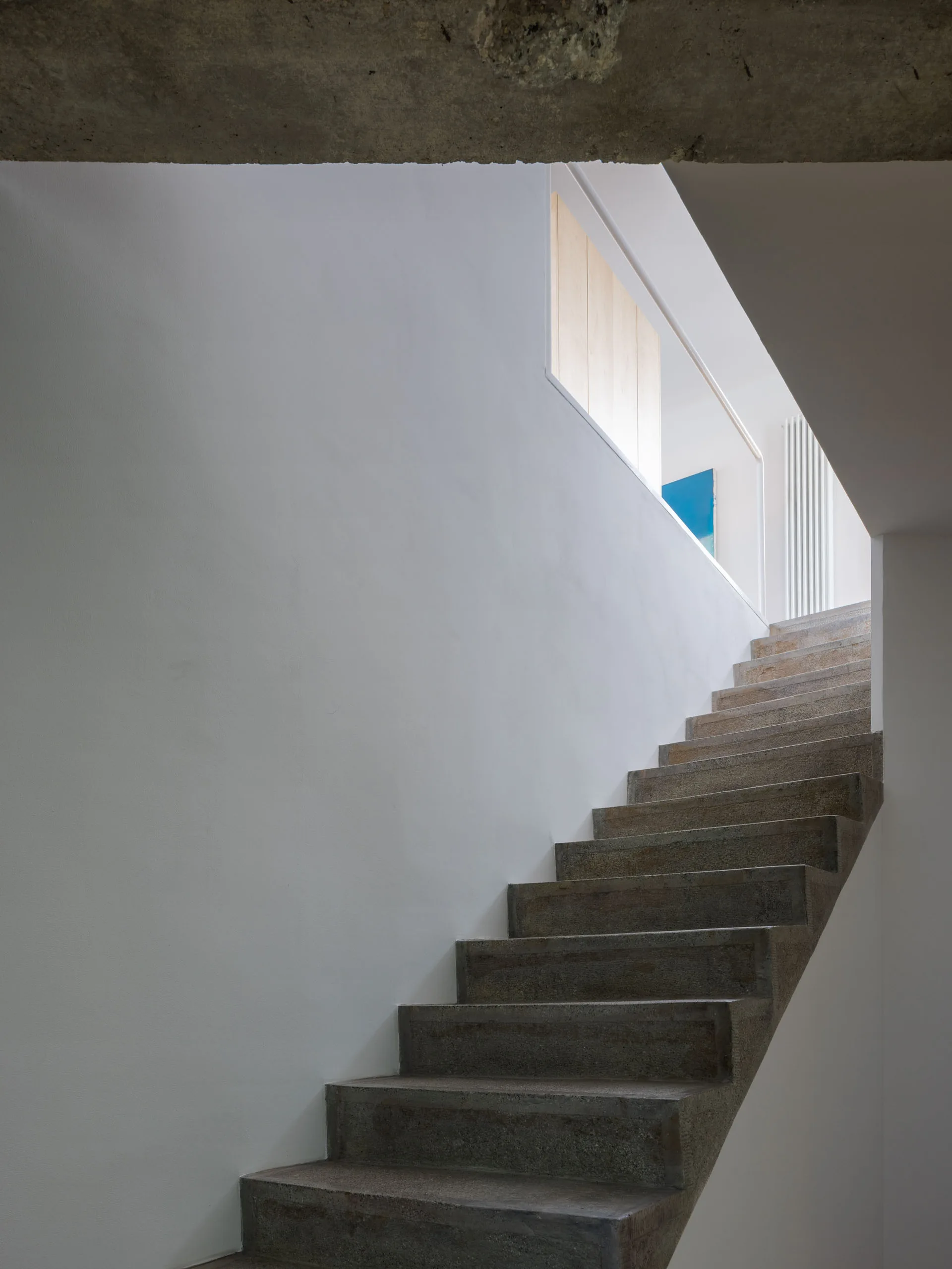 A minimalist staircase featuring grey concrete steps set against smooth white walls beneath an exposed concrete ceiling.