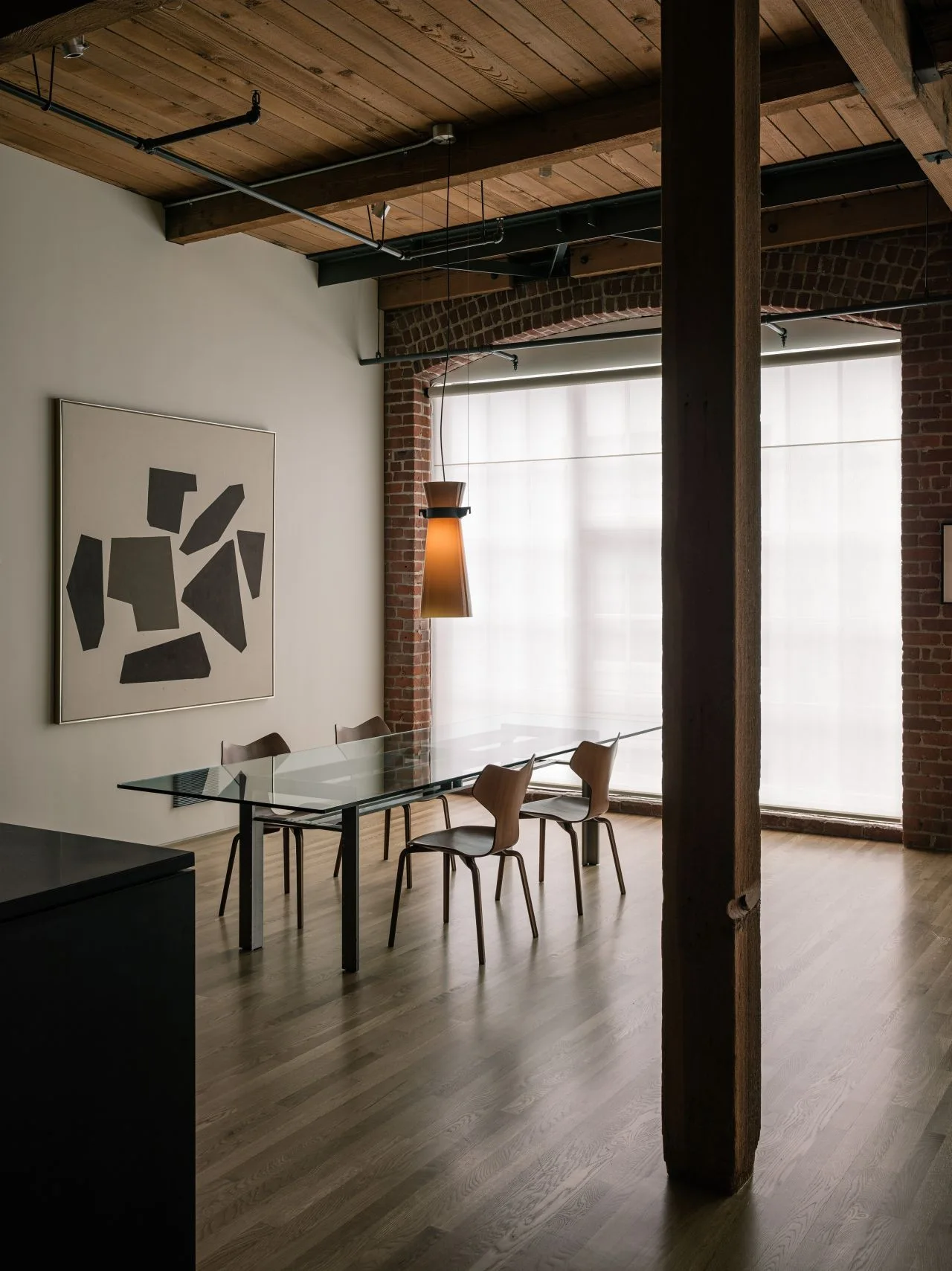 An industrial dining space features a glass table, wooden chairs, a large vertical column, and exposed brick walls under a timber ceiling.