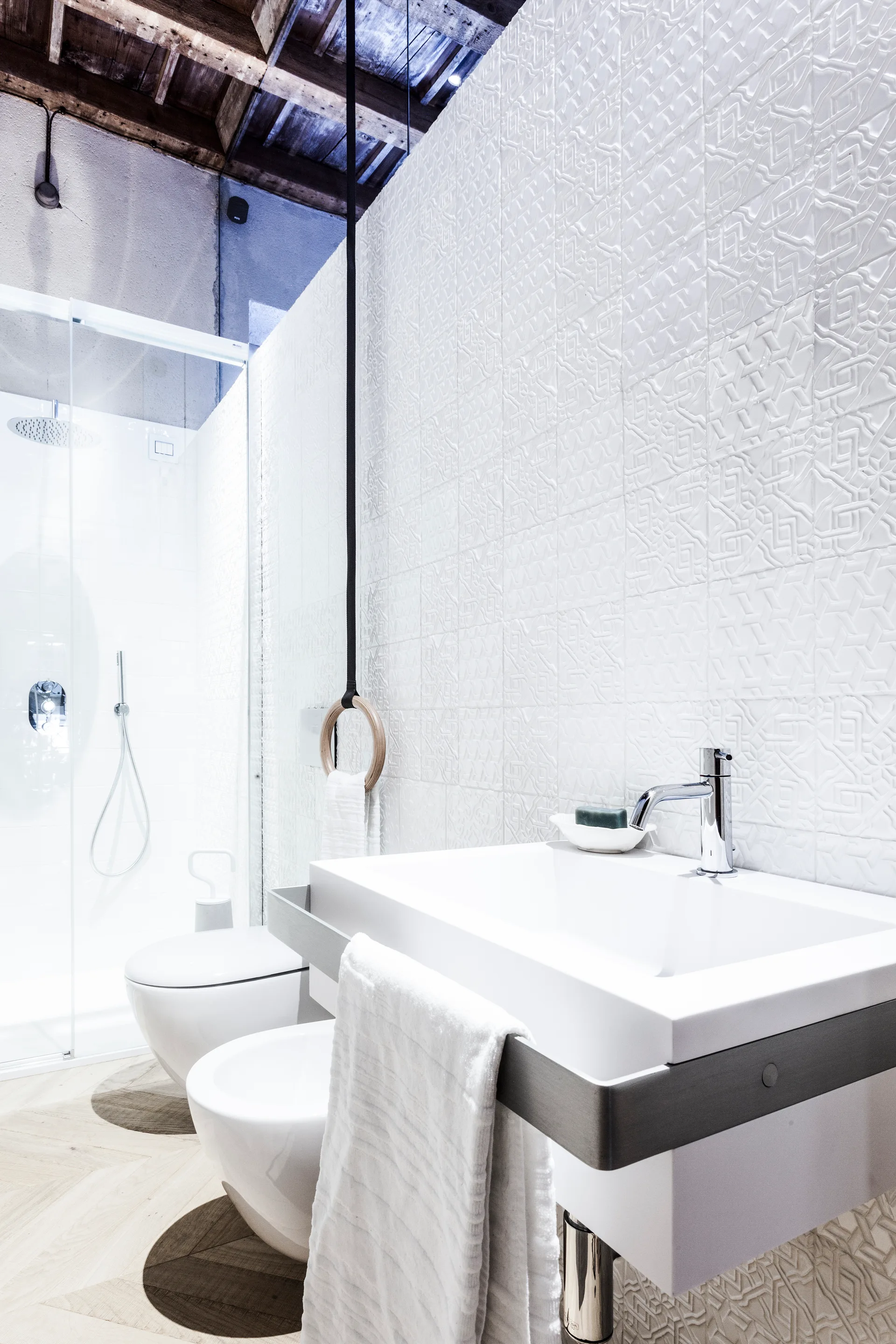 A modern bathroom features a white vanity sink against a geometric relief tile wall, with an exposed wood beam ceiling above.