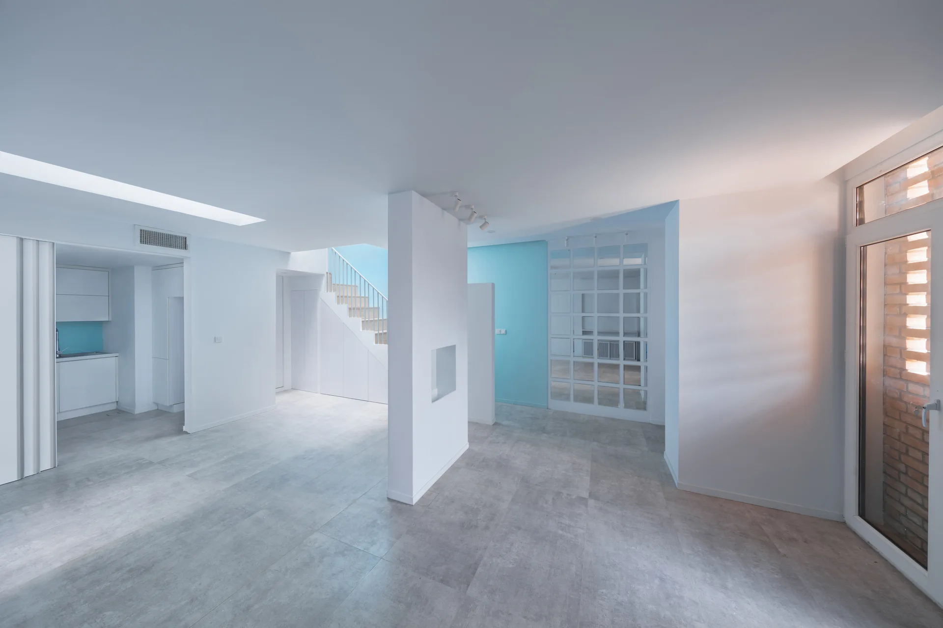 A minimalist open-plan living area features grey tiled flooring, white walls, a central partition column, and a light blue accent wall near a staircase.