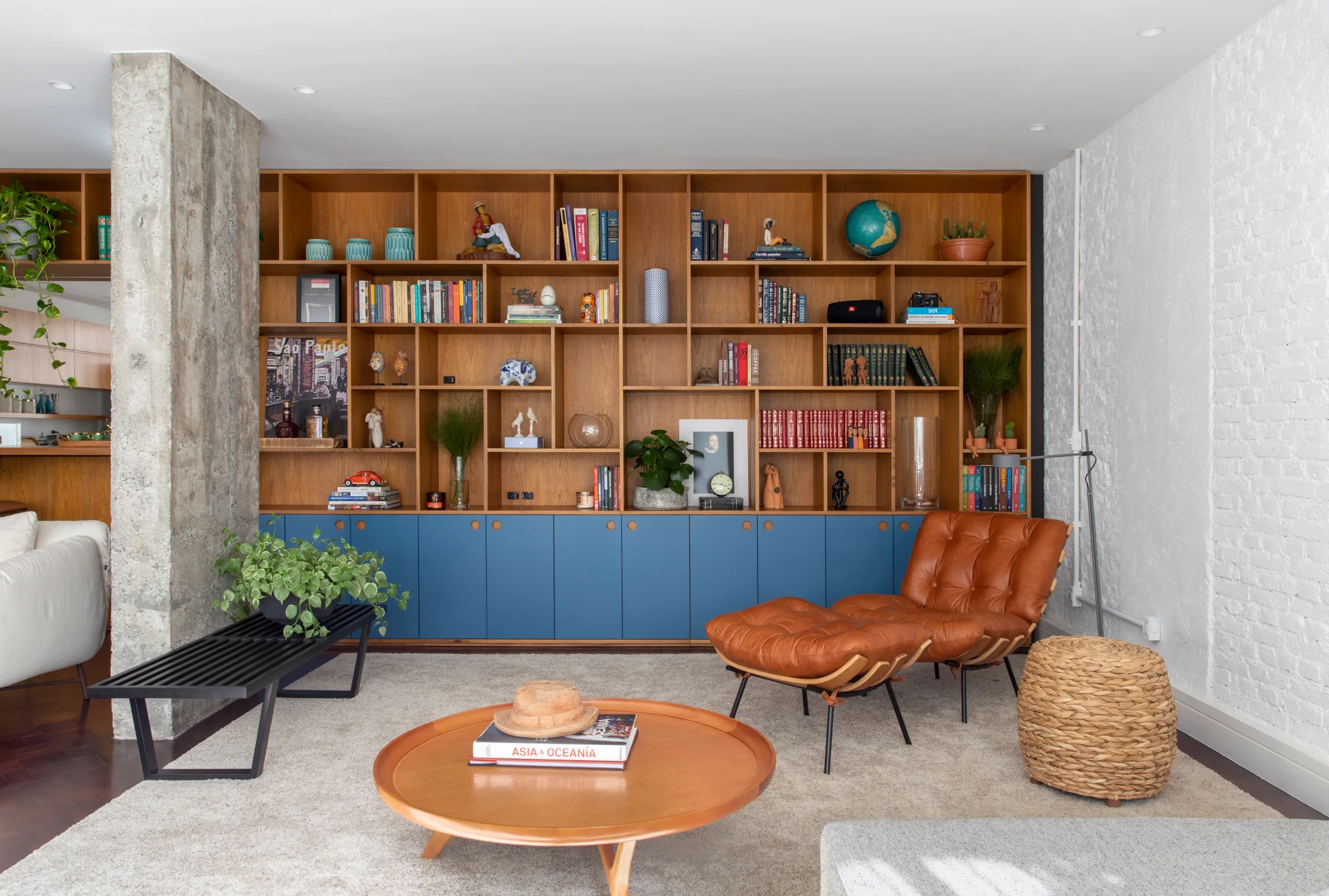 A living room features a tall wooden bookcase with blue lower doors, a brown leather lounge chair, and a concrete column on a beige rug.