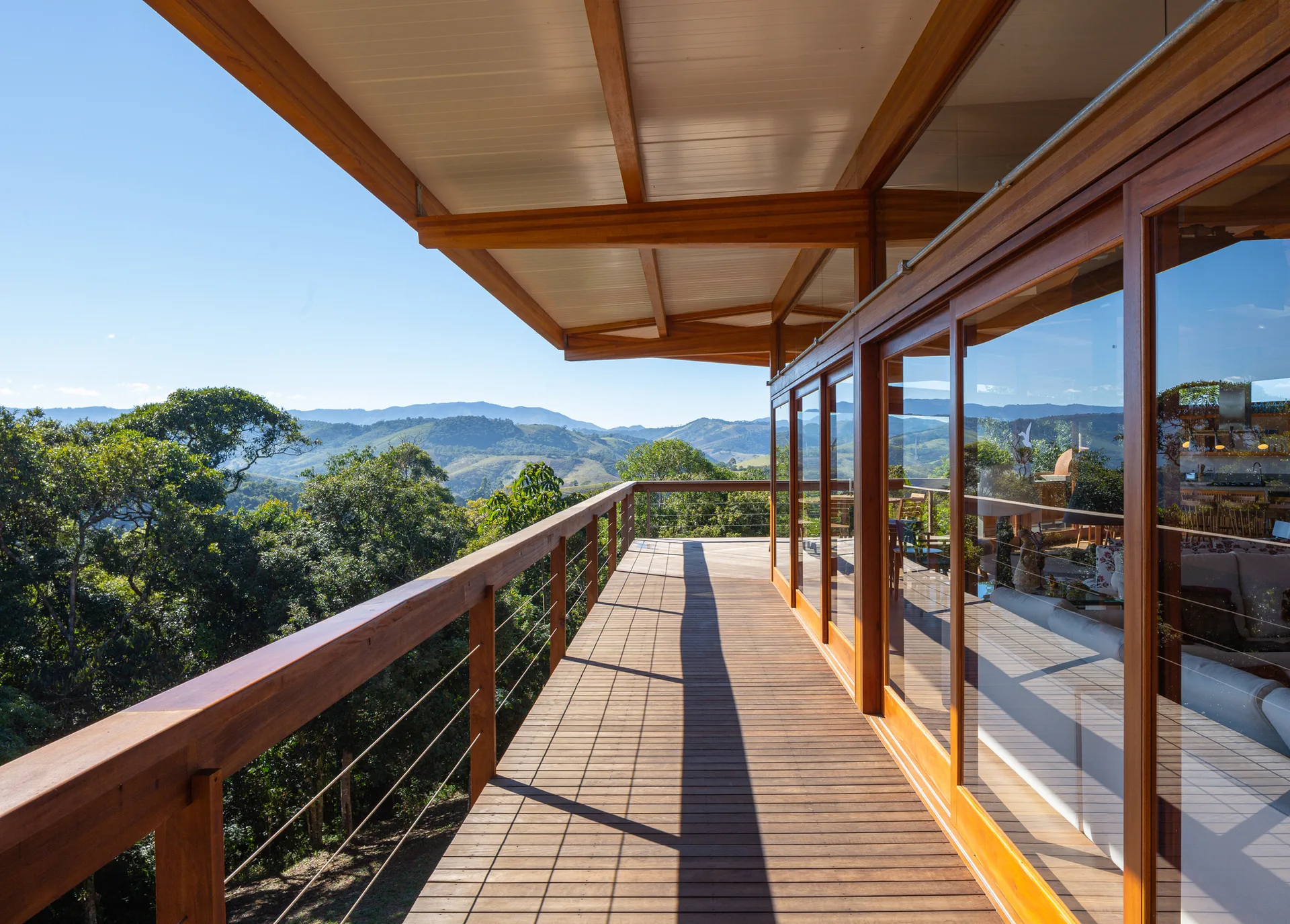 A long wooden terrace deck features a cable railing, sliding glass doors reflecting the interior, and a scenic view of green hills under a blue sky.