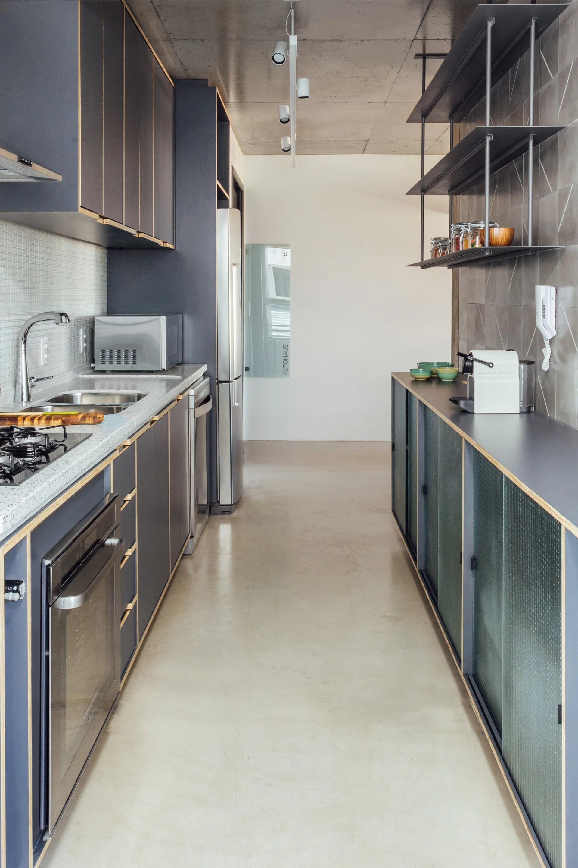 A galley kitchen featuring dark gray cabinets with exposed plywood edges, a raw concrete ceiling, and light gray concrete floors.