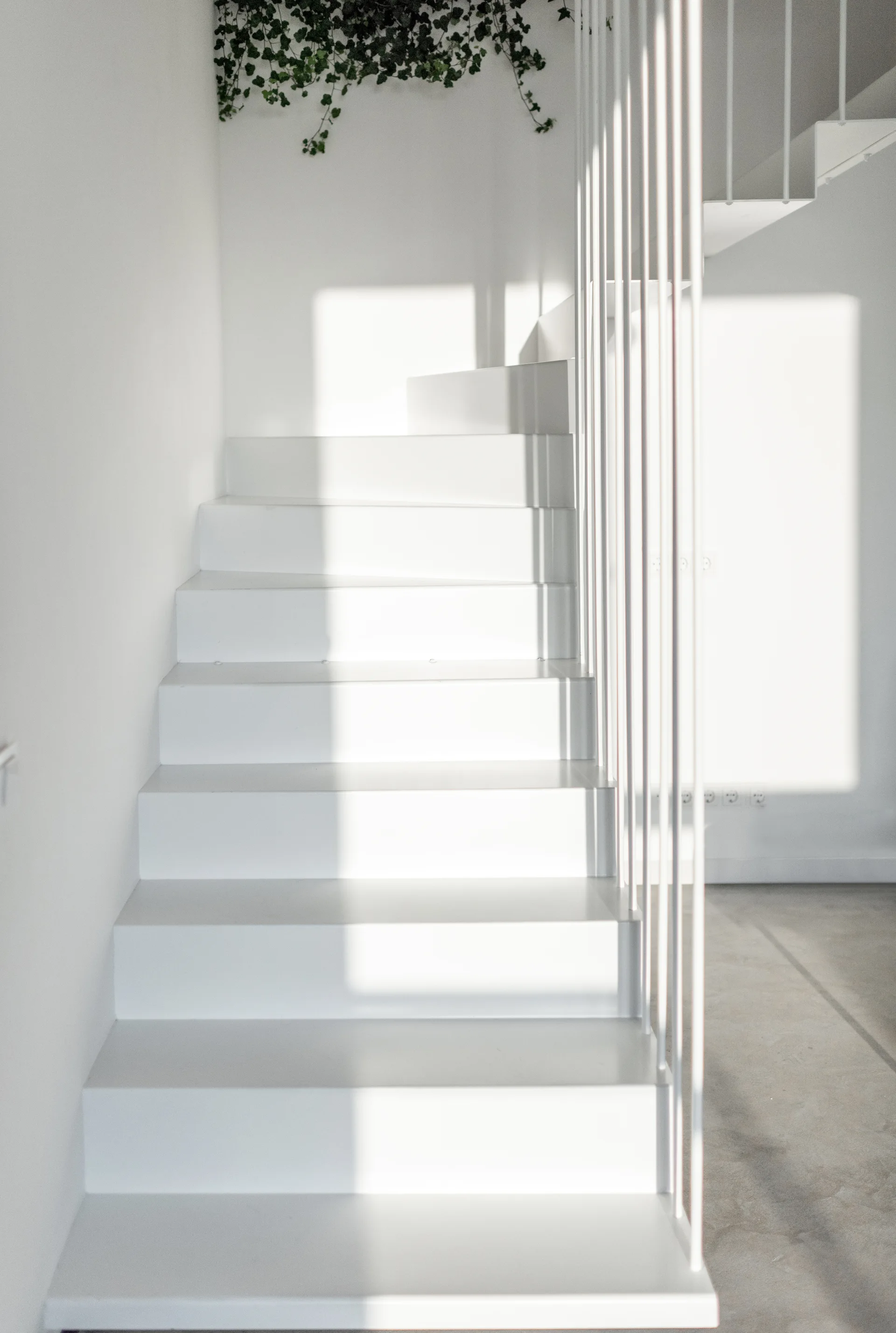 A minimalist white metal staircase featuring vertical rod railings and smooth treads, illuminated by sharp natural sunlight casting geometric shadows.