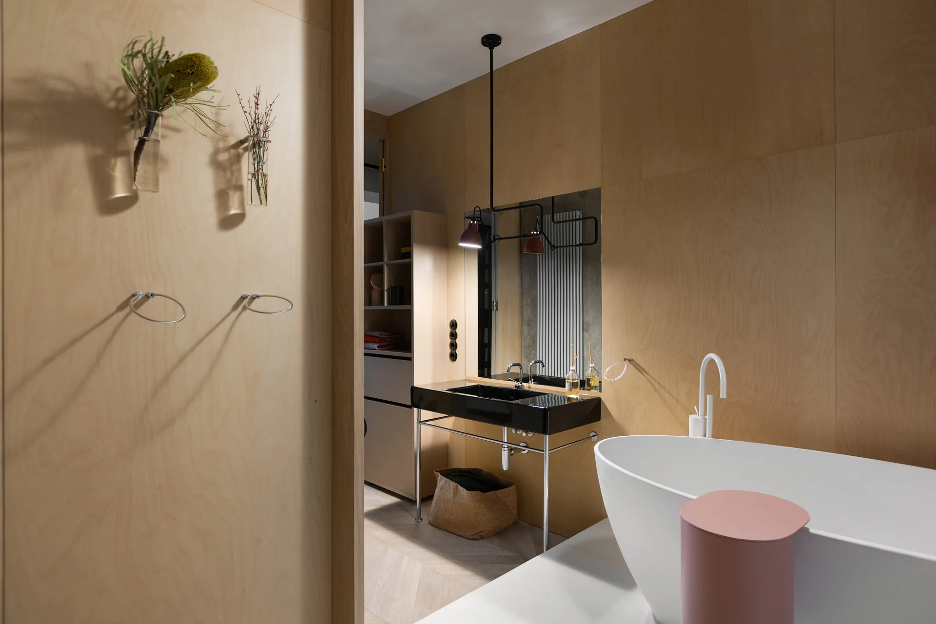 A modern bathroom with warm plywood walls features a white freestanding bathtub, a black console vanity with a mirror, and herringbone wood floors.