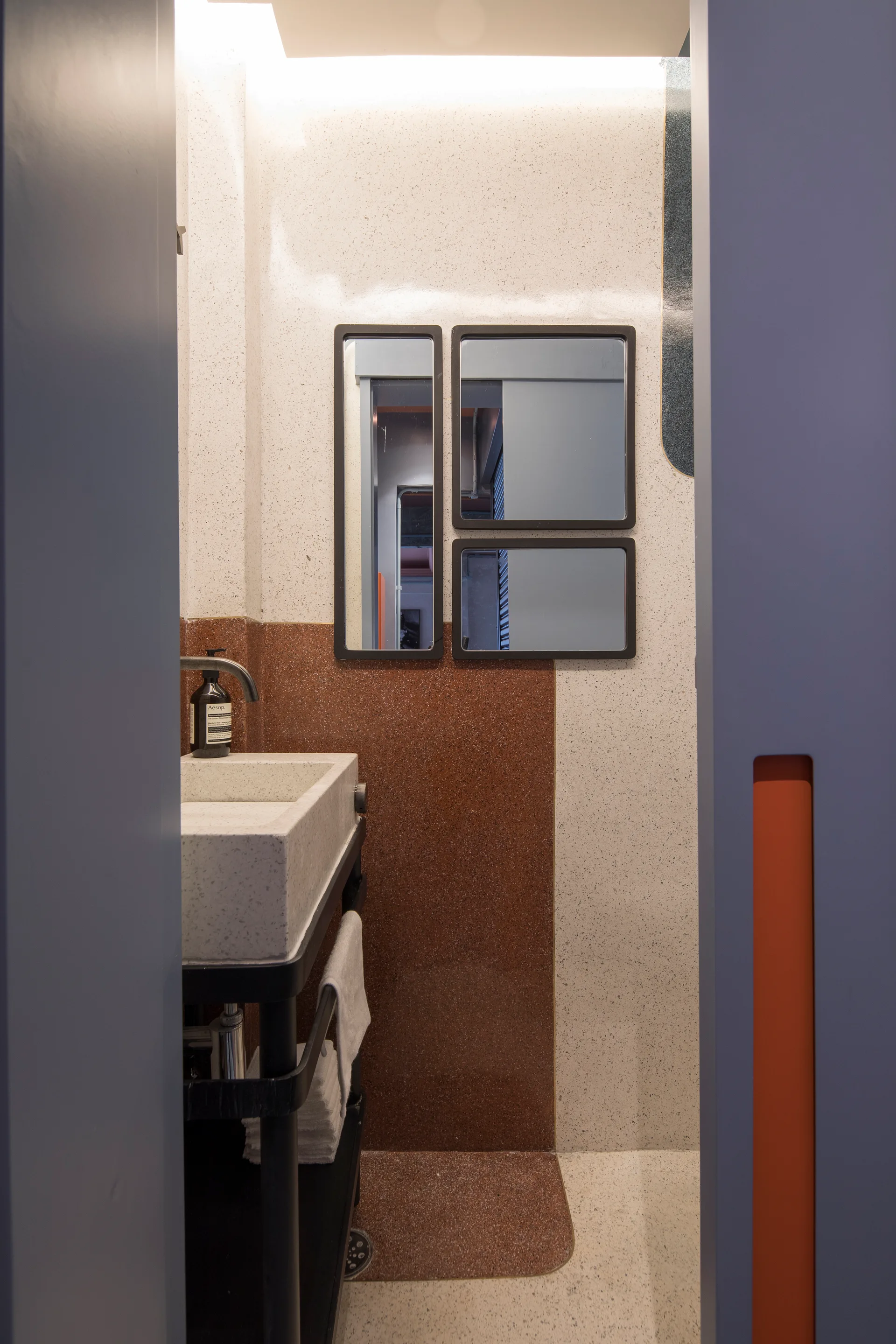 A modern bathroom features a speckled terrazzo wall, a rectangular stone basin sink, and three dark-framed rectangular mirrors.