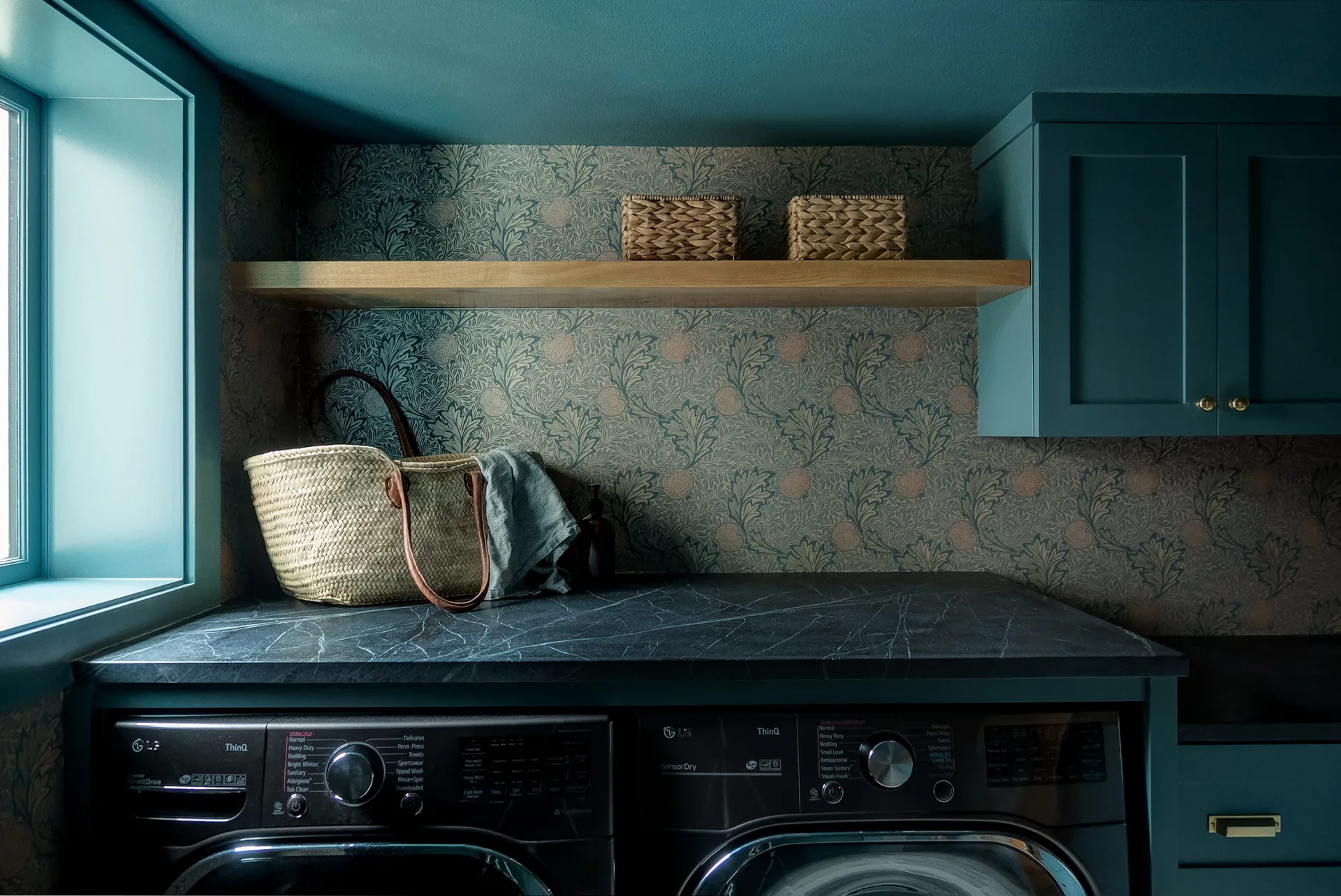 A laundry room features black laundry machines under a dark stone countertop, teal cabinets, botanical wallpaper, and a wooden shelf.