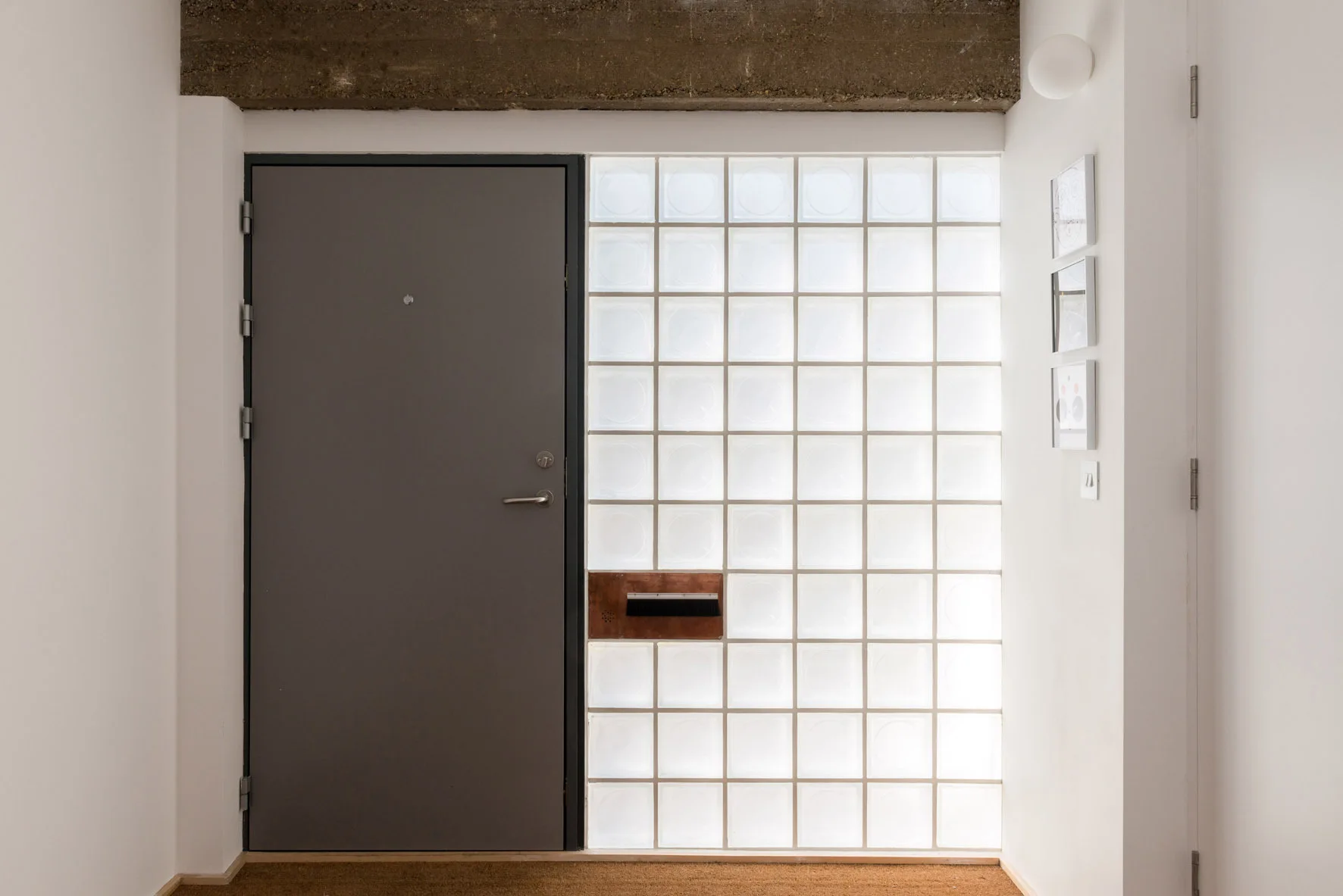 A minimalist entryway features a solid grey door, a translucent glass block wall, and an exposed concrete beam overhead.