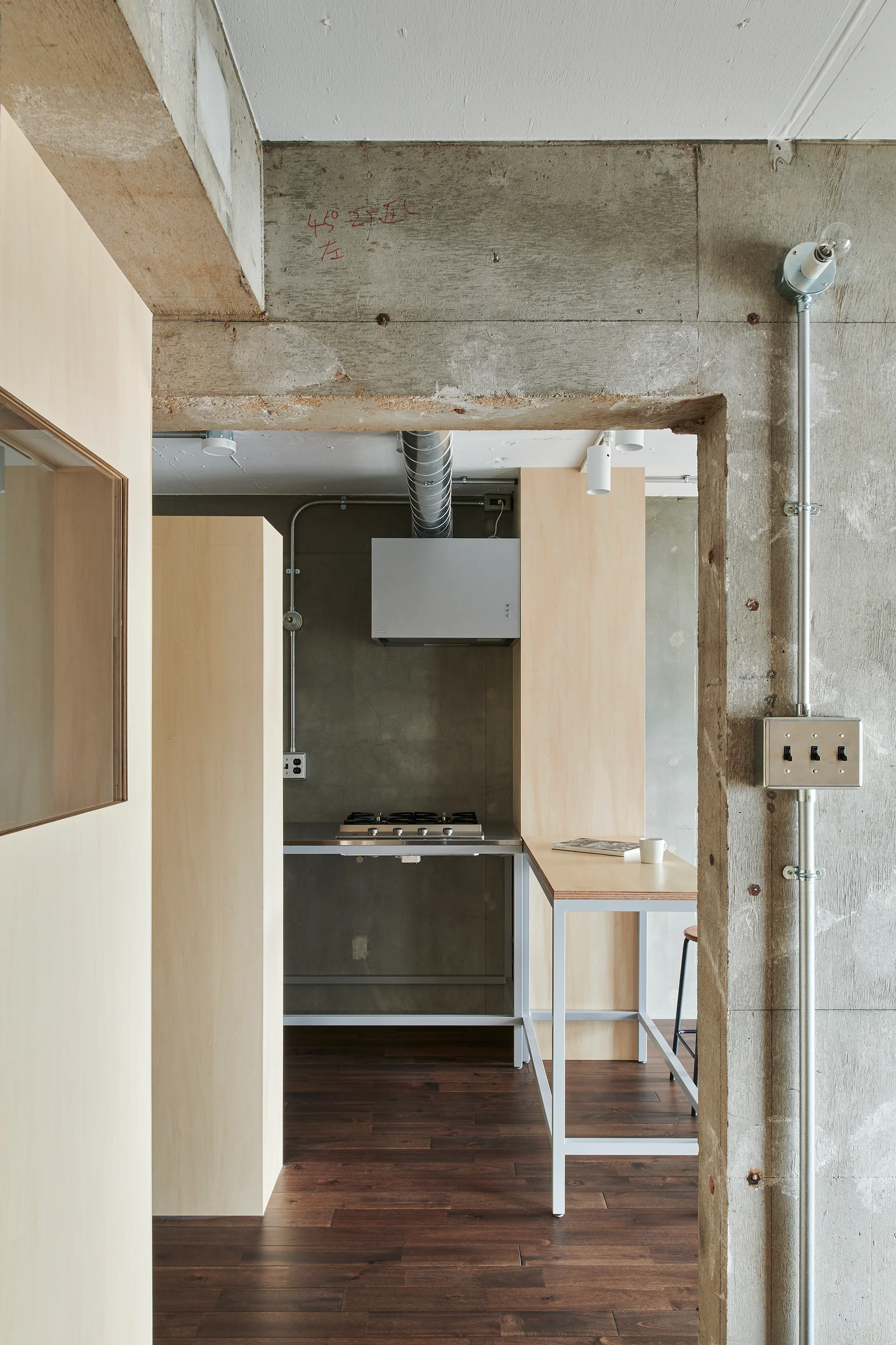A view through a concrete doorway into an industrial kitchen with raw concrete walls, light plywood cabinets, a metal stovetop, and dark wood flooring.