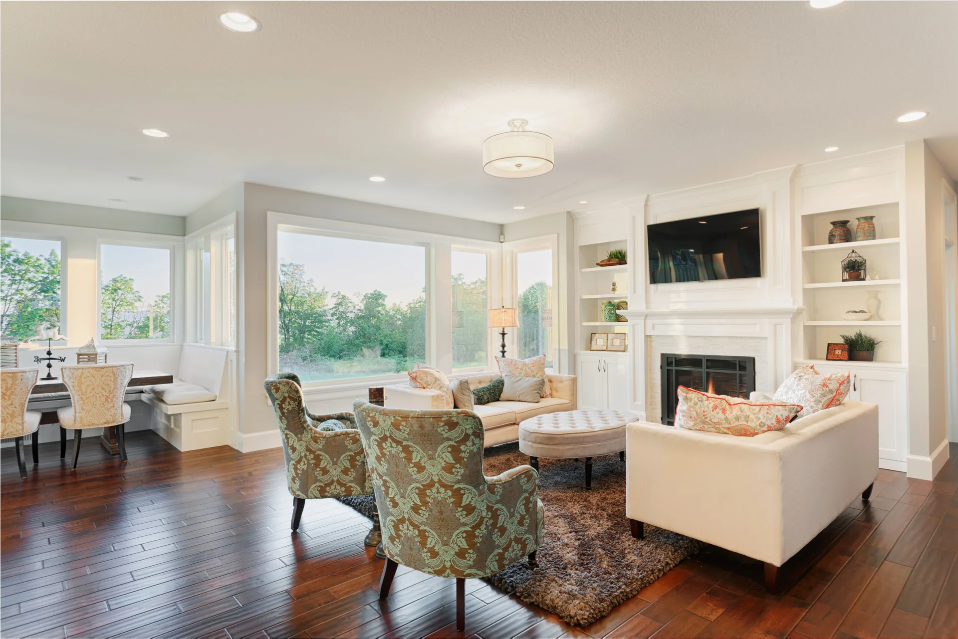 A bright living room featuring a white fireplace flanked by custom built-in shelving, cream sofas, and patterned armchairs on dark hardwood floors.