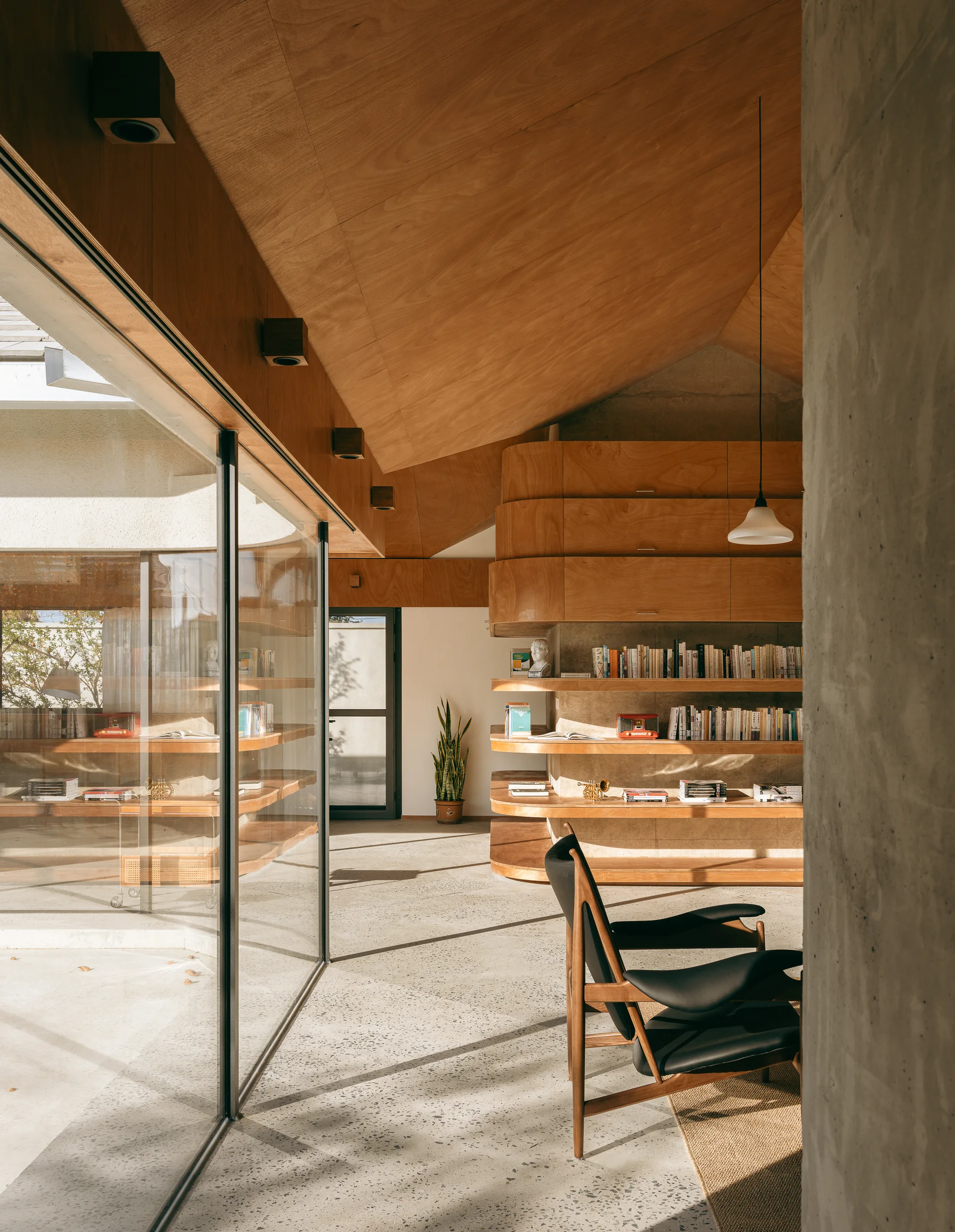 A modern room featuring a vaulted wood panel ceiling, curved plywood shelving with books, a black armchair, and a terrazzo-style concrete floor.