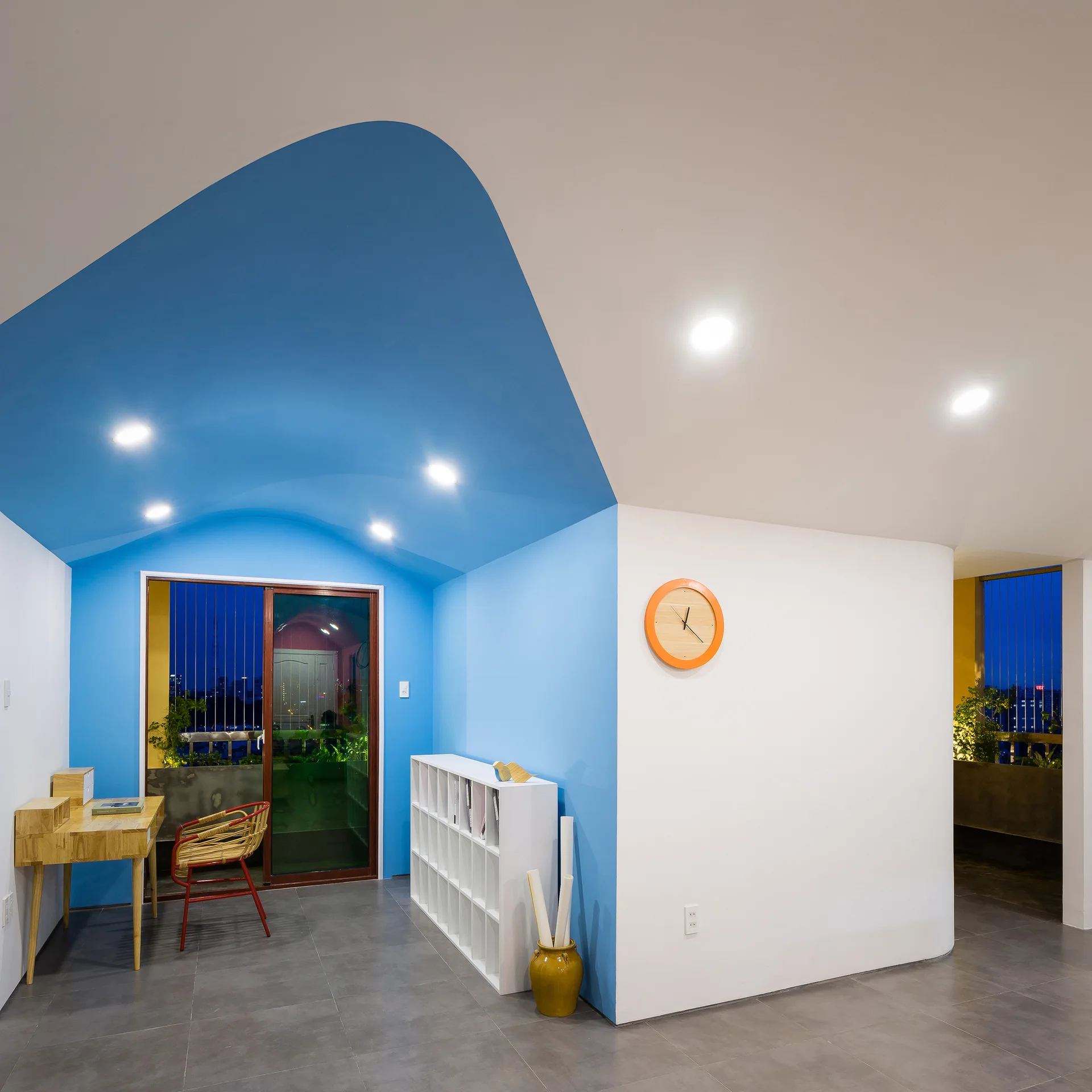 A home office features a bright blue painted vaulted ceiling segment above a wooden desk, white cubby shelf, and grey tiled flooring.