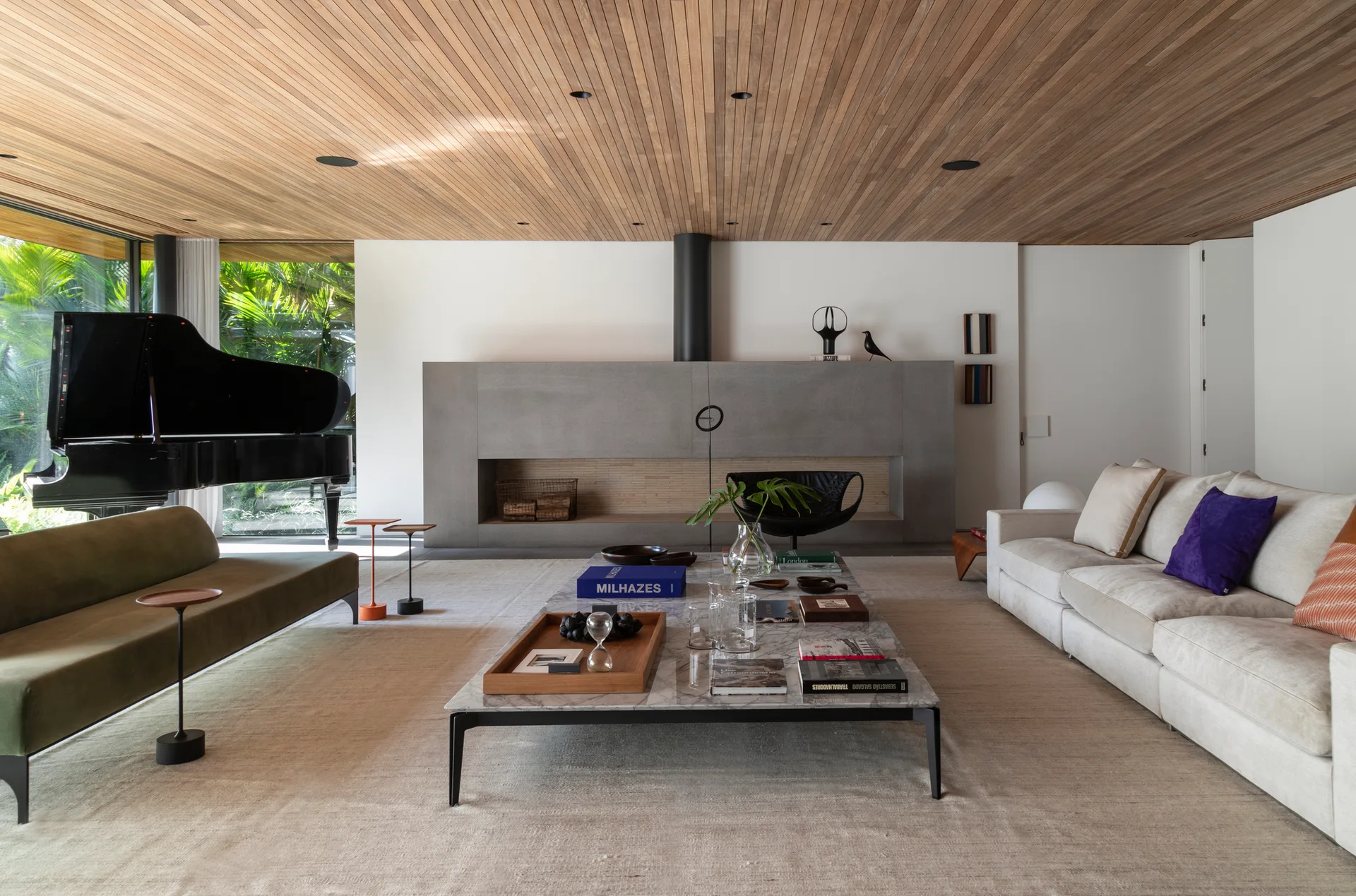 A spacious living room features a slatted wood ceiling, a large grey stone fireplace, a grand piano, and a cream sofa.