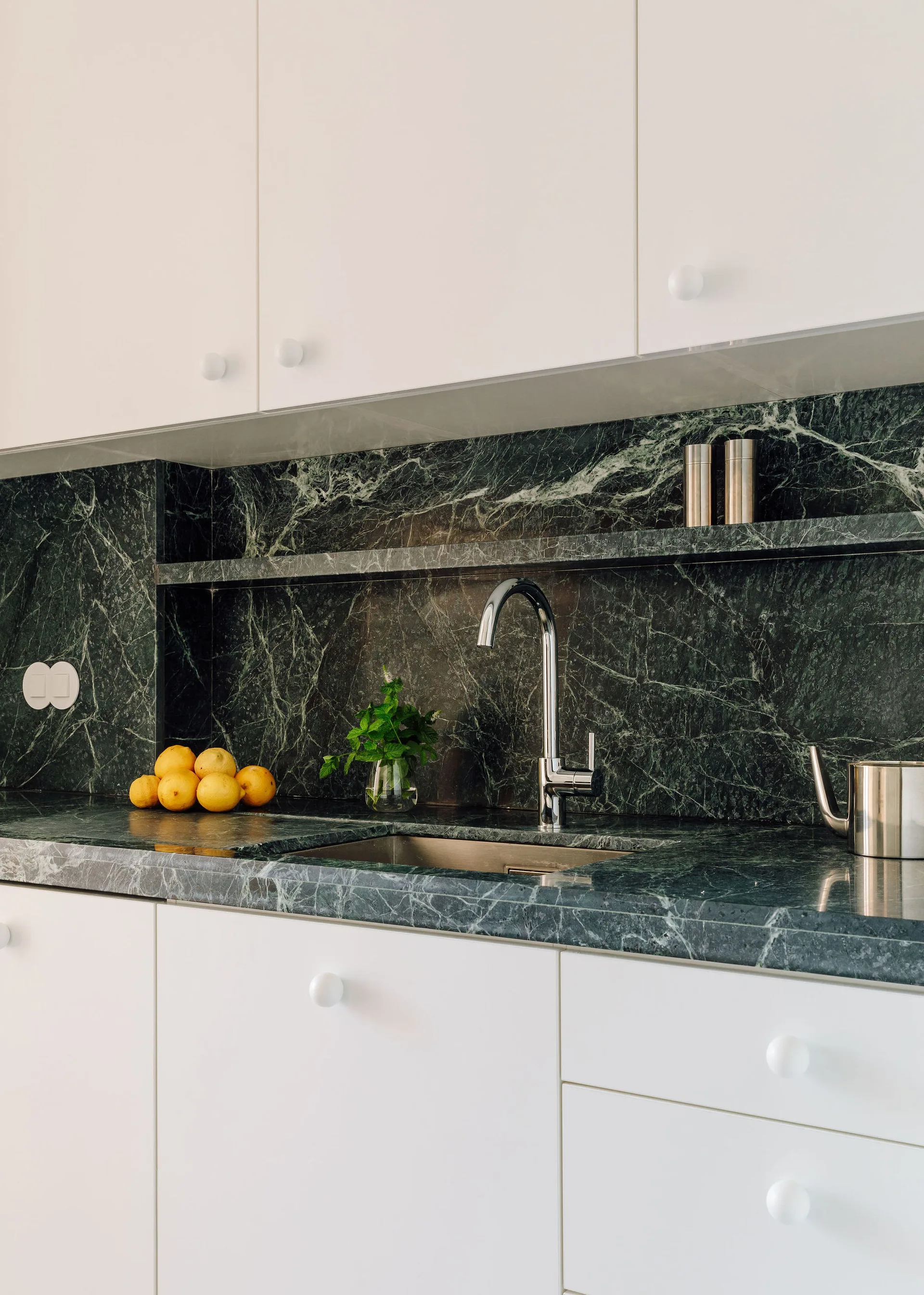 A minimalist kitchen sink with white cabinetry, a dark green marble countertop, and a matching stone slab backsplash with a built-in ledge.