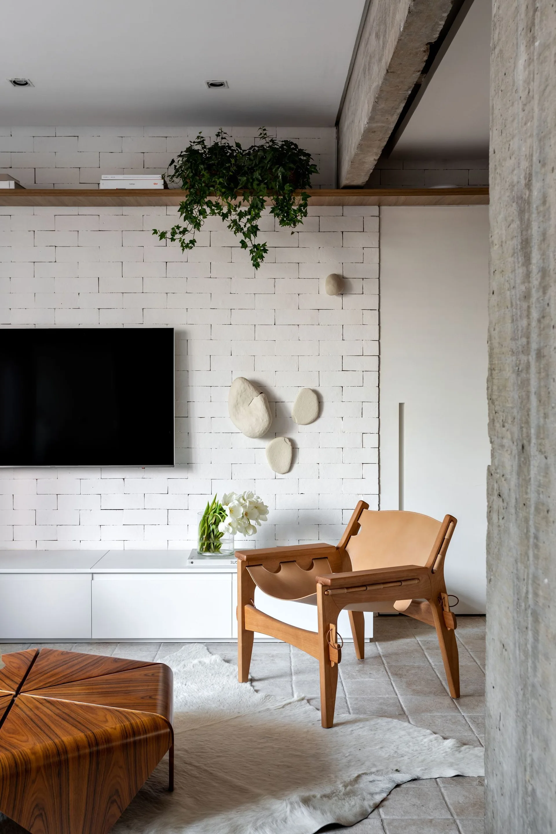 A modern living room features a tan leather lounge chair, a low white console against a painted white brick wall, and a geometric wood coffee table.