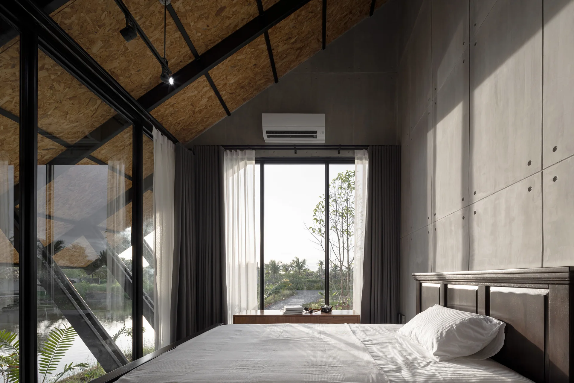 A modern industrial bedroom features a bed with white linens next to a concrete panel wall beneath a pitched OSB ceiling and large windows.