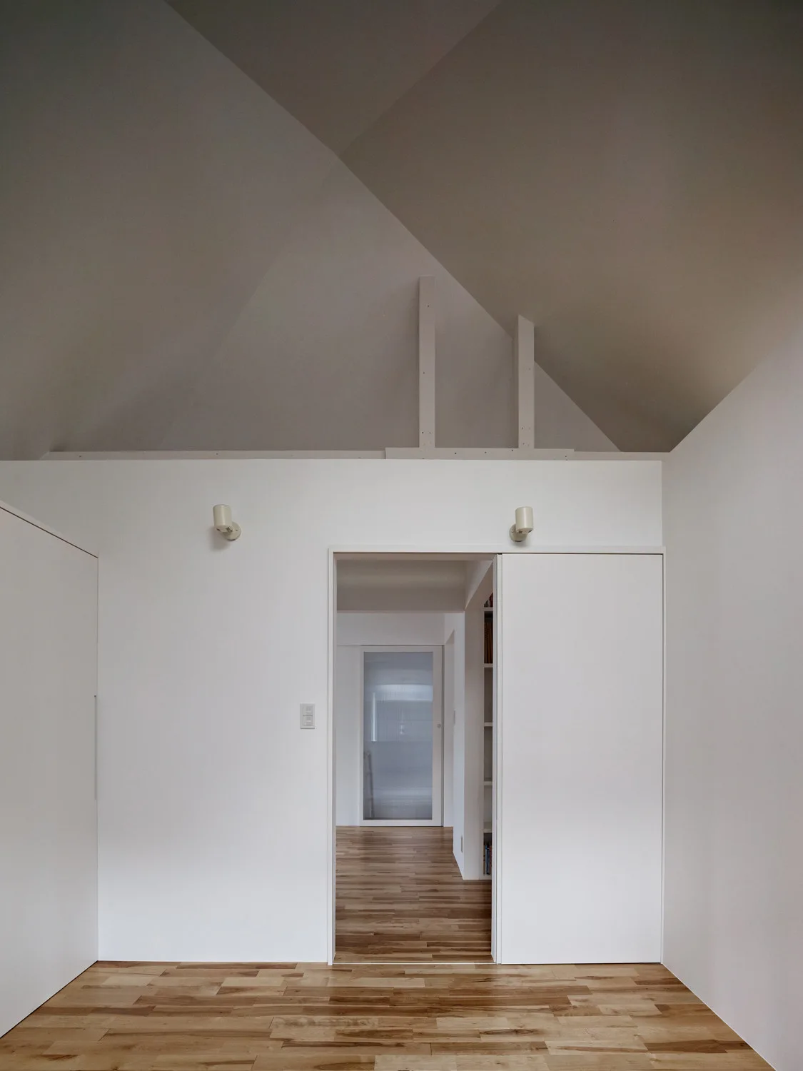 A minimalist hallway features matte white walls, a vaulted ceiling, wood plank flooring, and a wide open doorway with a sliding white door panel.