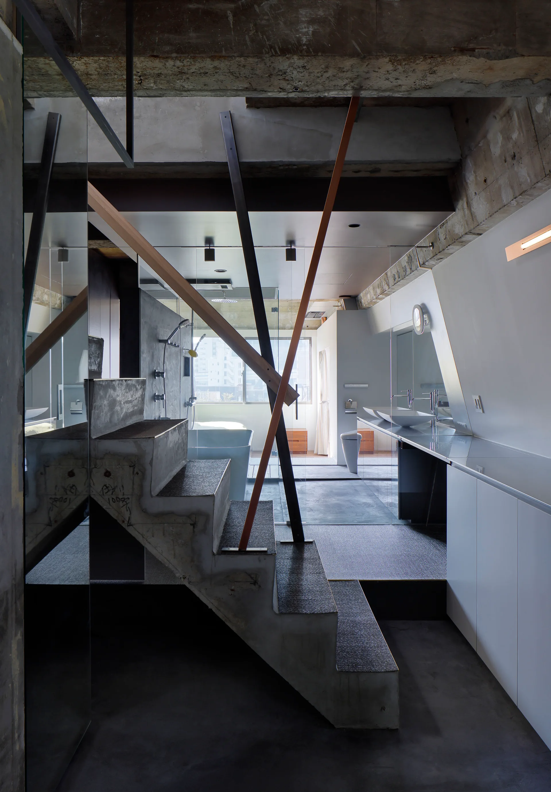 An industrial bathroom showing a central raw concrete staircase beside a long white vanity with a vessel sink and glass partitions.