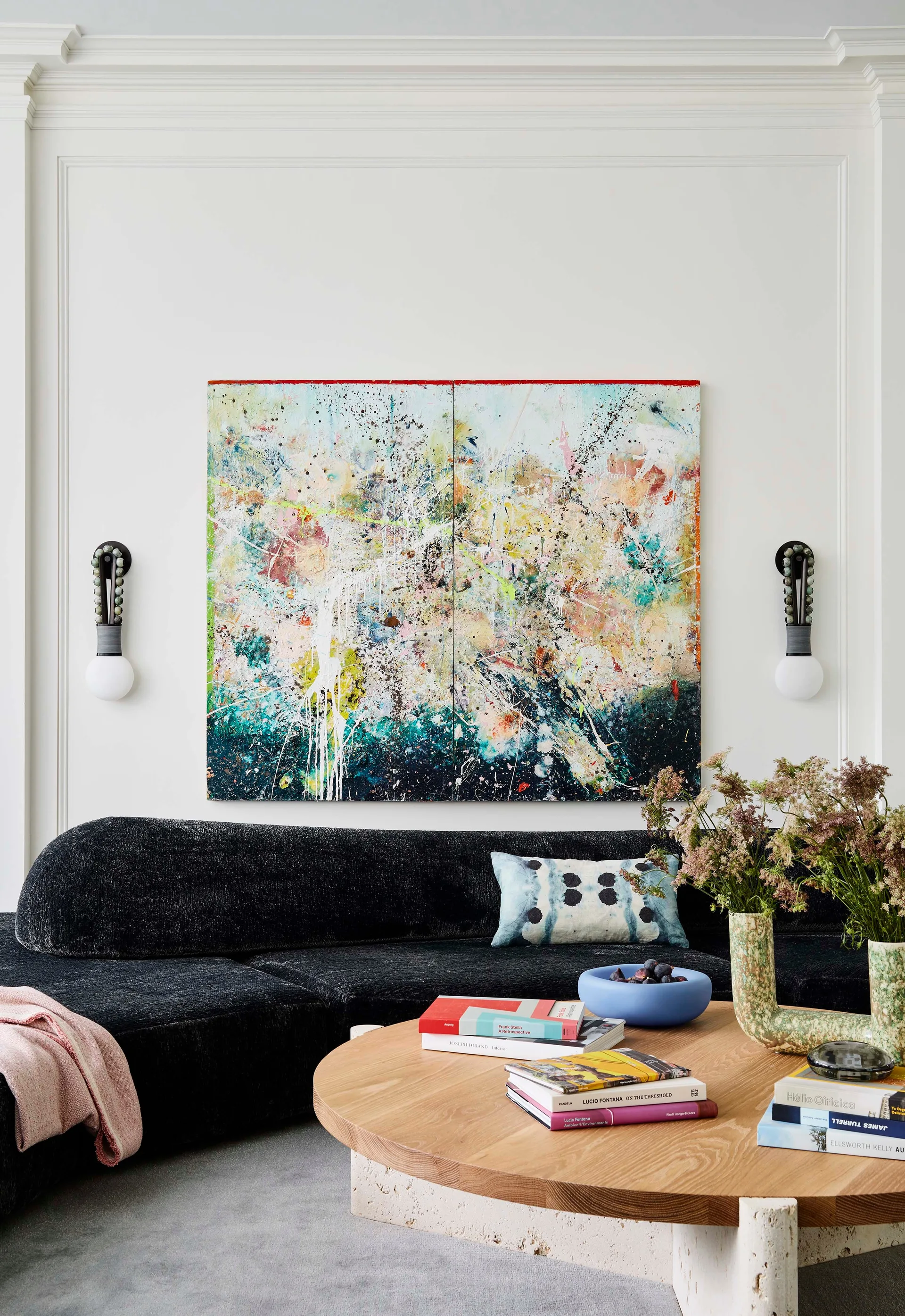 A large abstract painting is flanked by modern black and white wall sconces on a white paneled wall above a dark textured sofa.