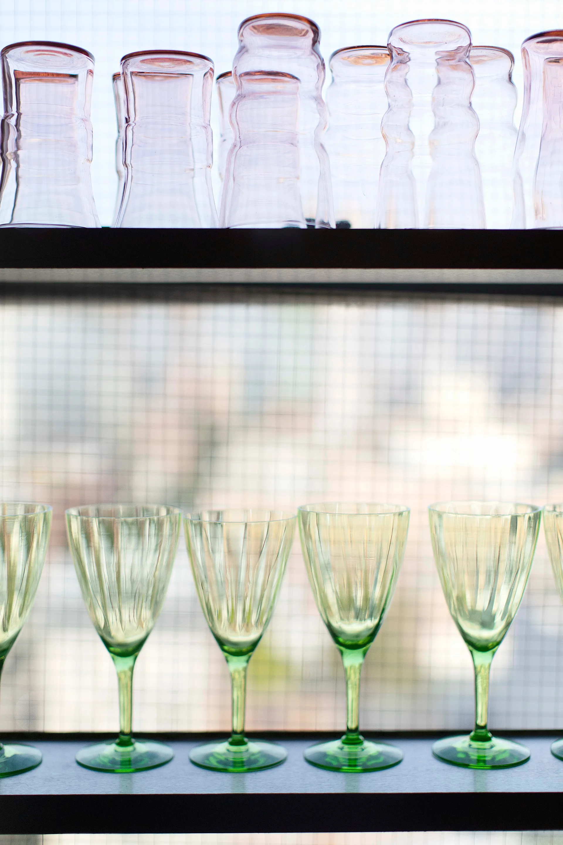 Pink tumblers and green stemmed wine glasses sit on dark open shelves backlit by a grid-patterned window.