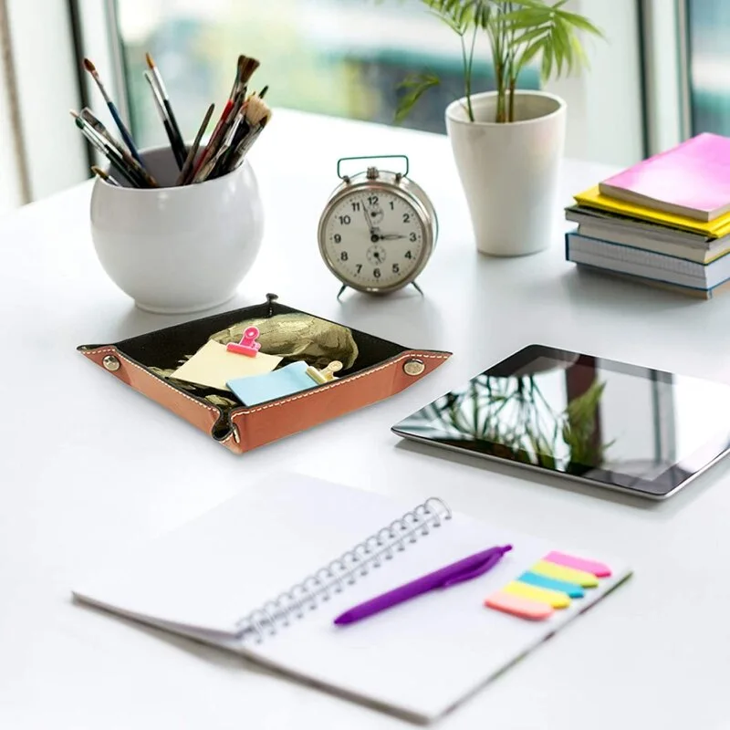 A brown leather valet tray with office accessories sits on a bright white desk alongside a notebook, tablet, and vintage alarm clock.