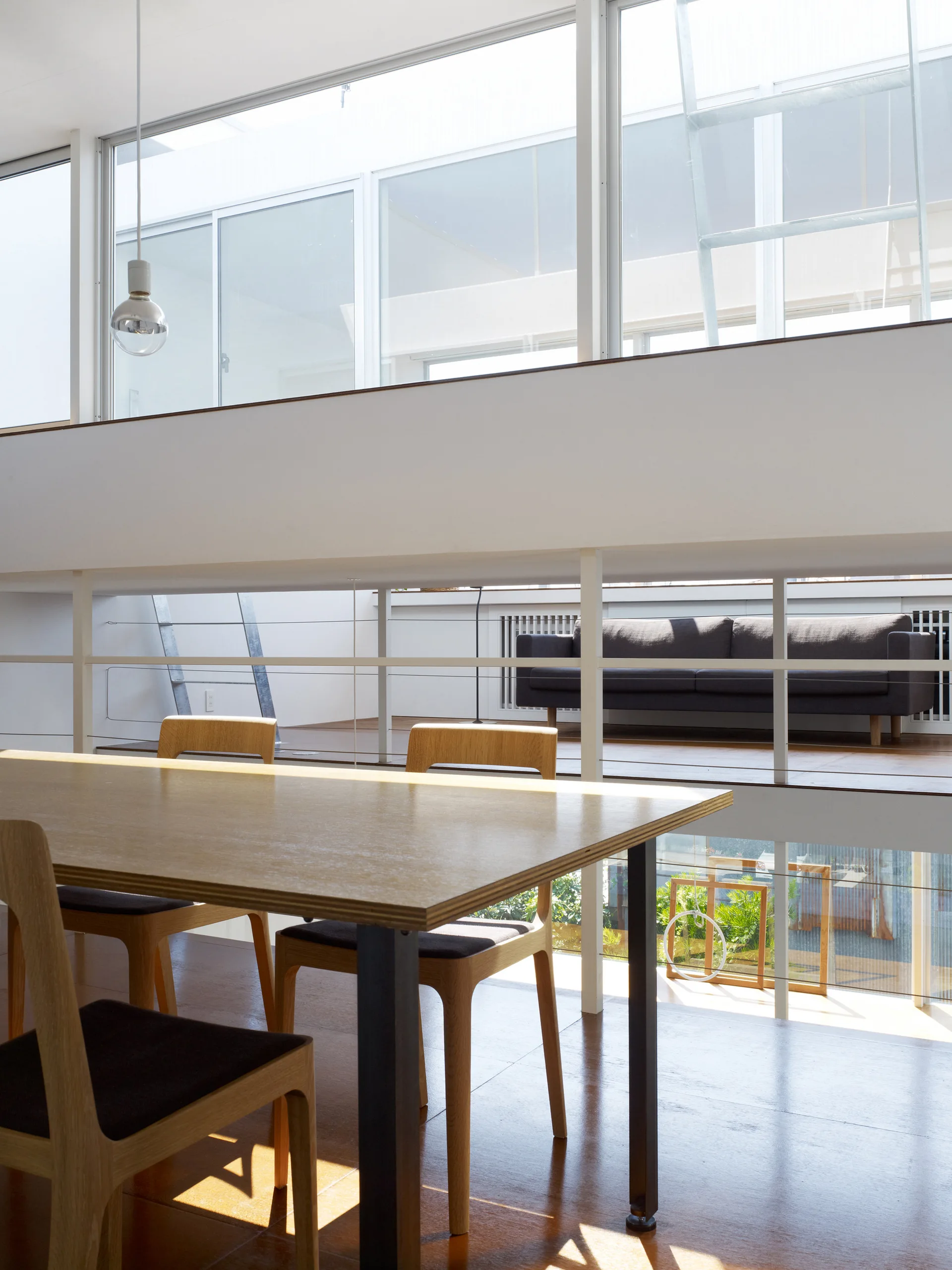 A modern split-level dining area features a wood dining table and chairs in the foreground, with a grey sofa visible on the mezzanine level above.