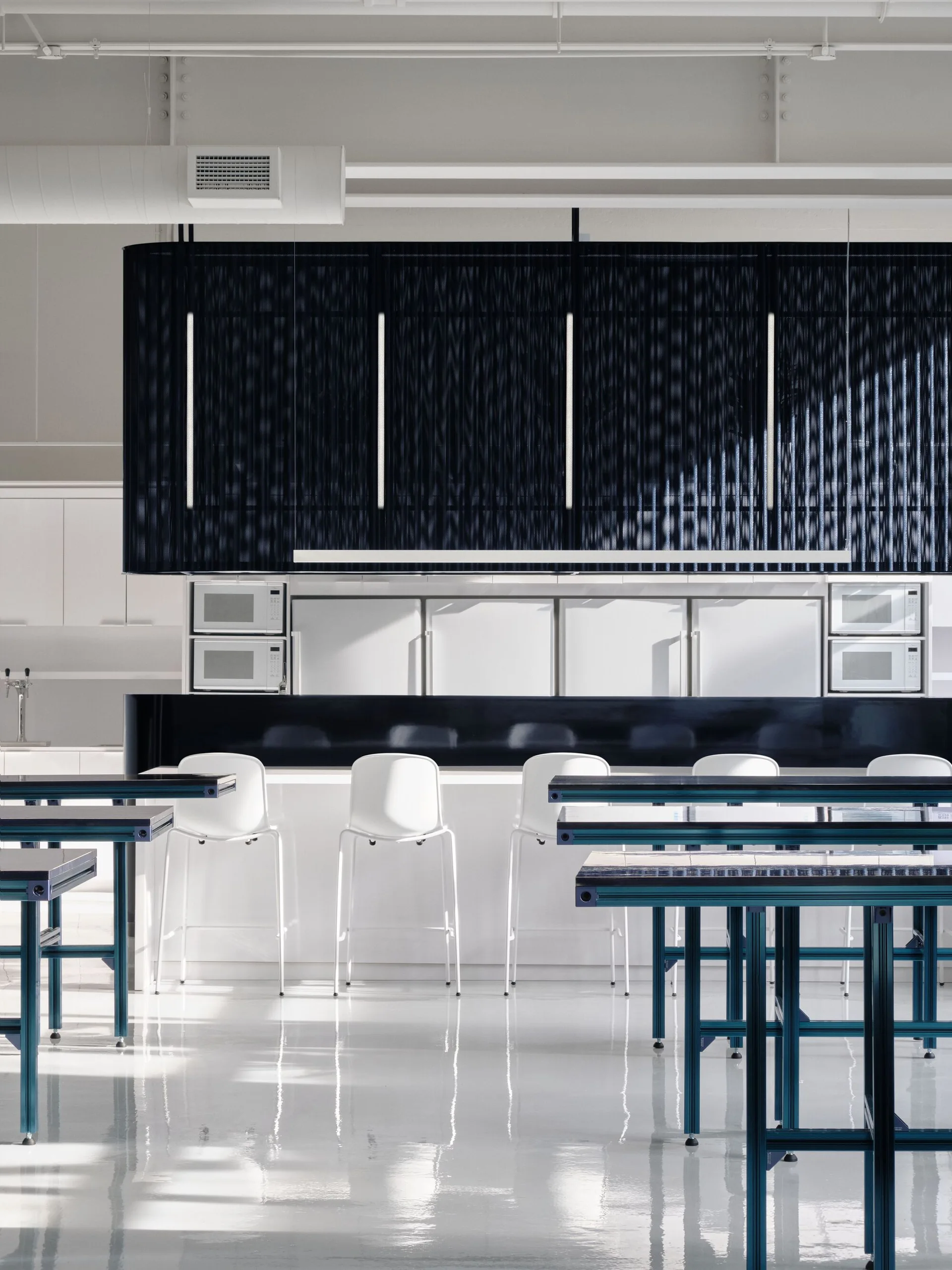 A modern industrial kitchen features white bar stools at a white island beneath a large navy blue hanging canopy and exposed white ceiling pipes.
