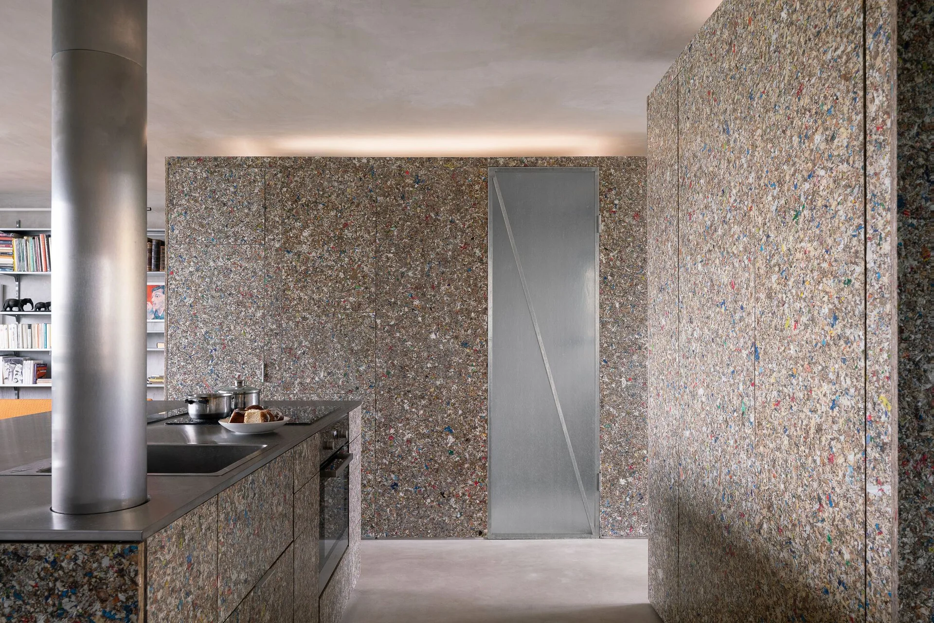 A modern kitchen featuring a stainless steel island and cabinets clad in speckled multicolor recycled composite panels beneath a concrete ceiling.