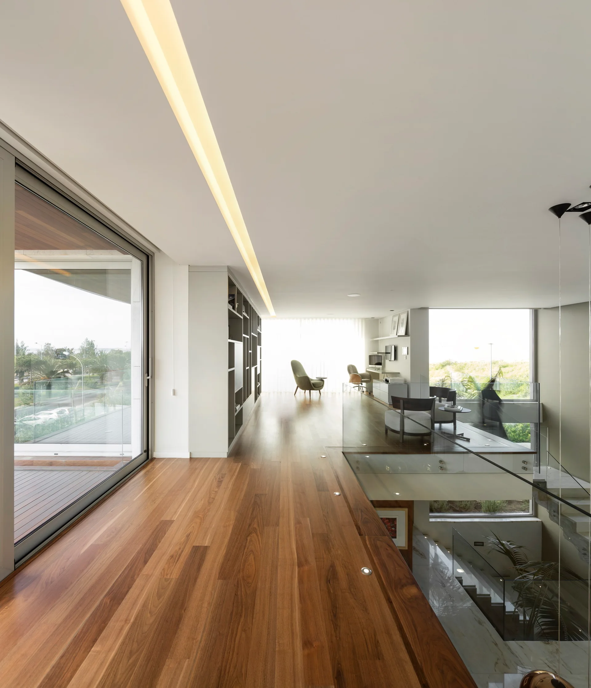 A minimalist mezzanine landing features warm hardwood flooring, a glass railing overlooking the lower floor, and a linear recessed ceiling light.