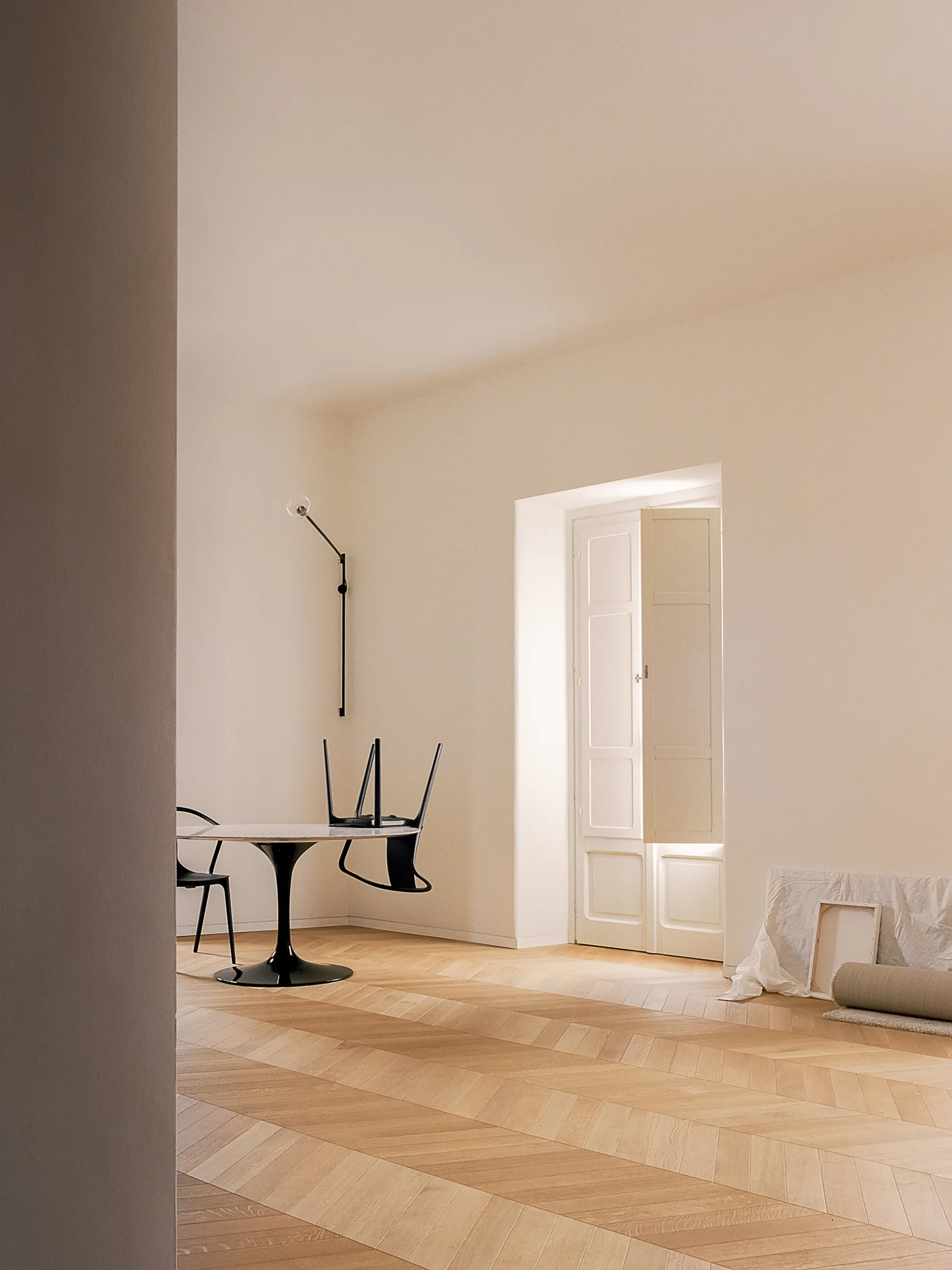 A minimalist dining room features a black pedestal table and chairs on a light wood herringbone floor near a white paneled double door.