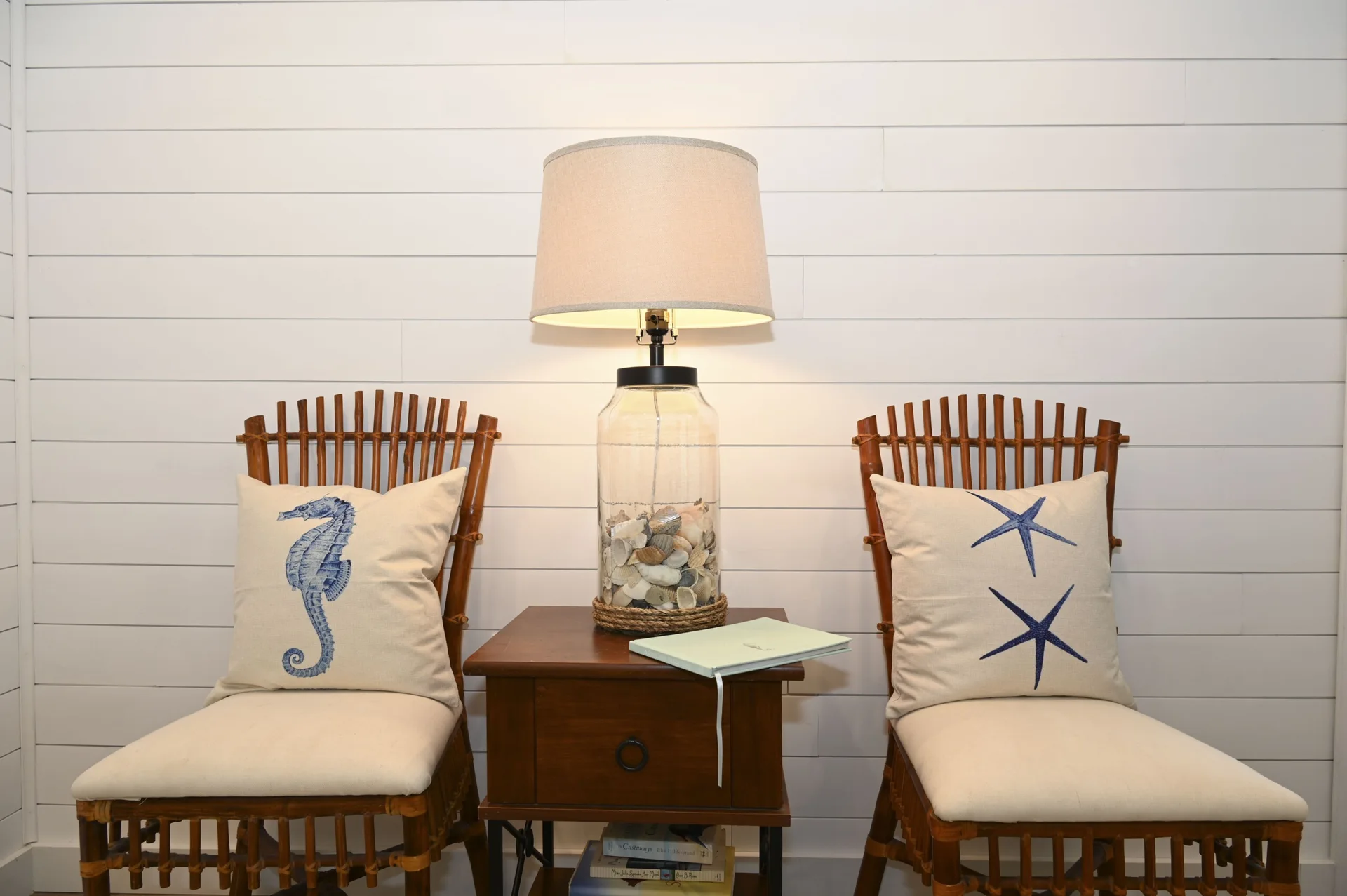 Two brown bamboo chairs with nautical throw pillows flank a wooden side table and a glass jar shell lamp against a white shiplap wall.