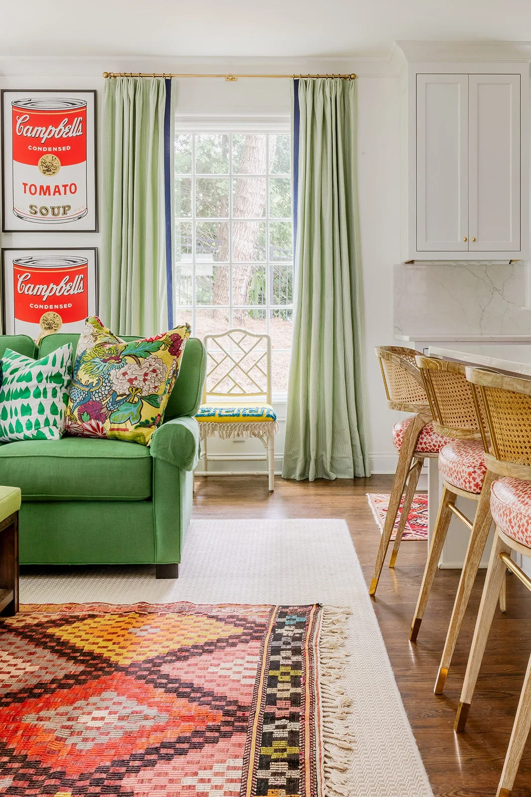A green sofa with patterned pillows sits next to a window with light green curtains, near cane-back bar stools and a geometric rug.