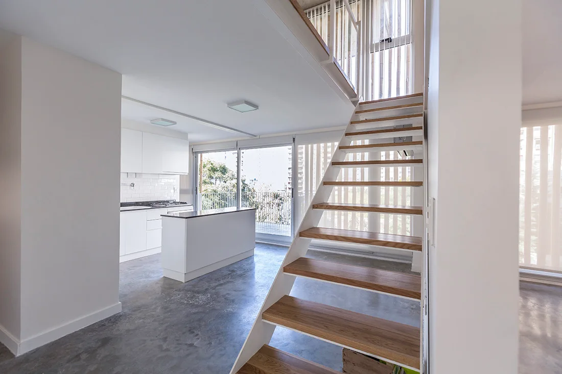 An open-riser wooden staircase stands in a modern loft next to a white minimalist kitchen and a polished grey concrete floor.