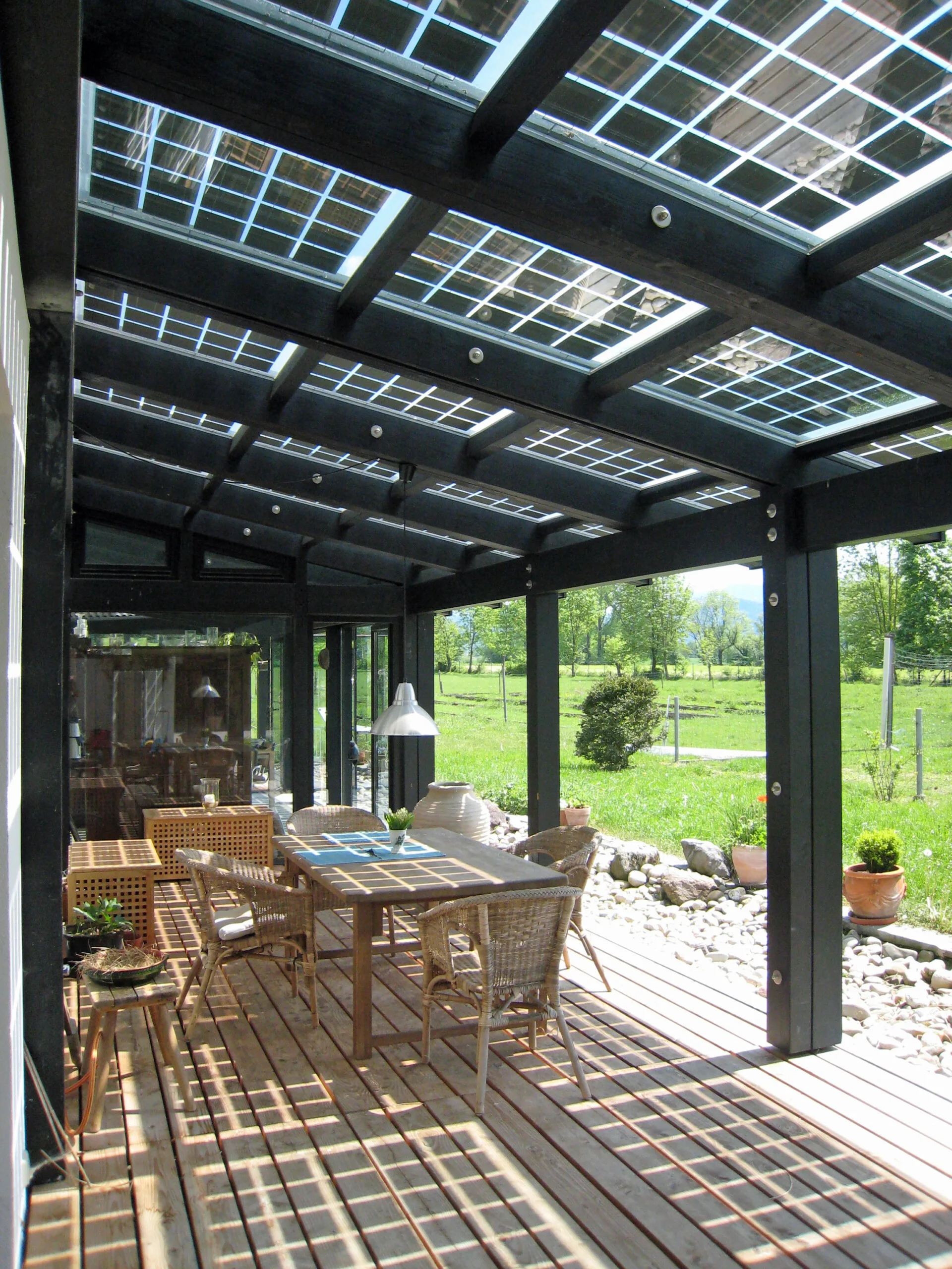 A covered wooden deck patio featuring a dining set under a black-framed glass pergola roof with integrated solar panels next to a green lawn.