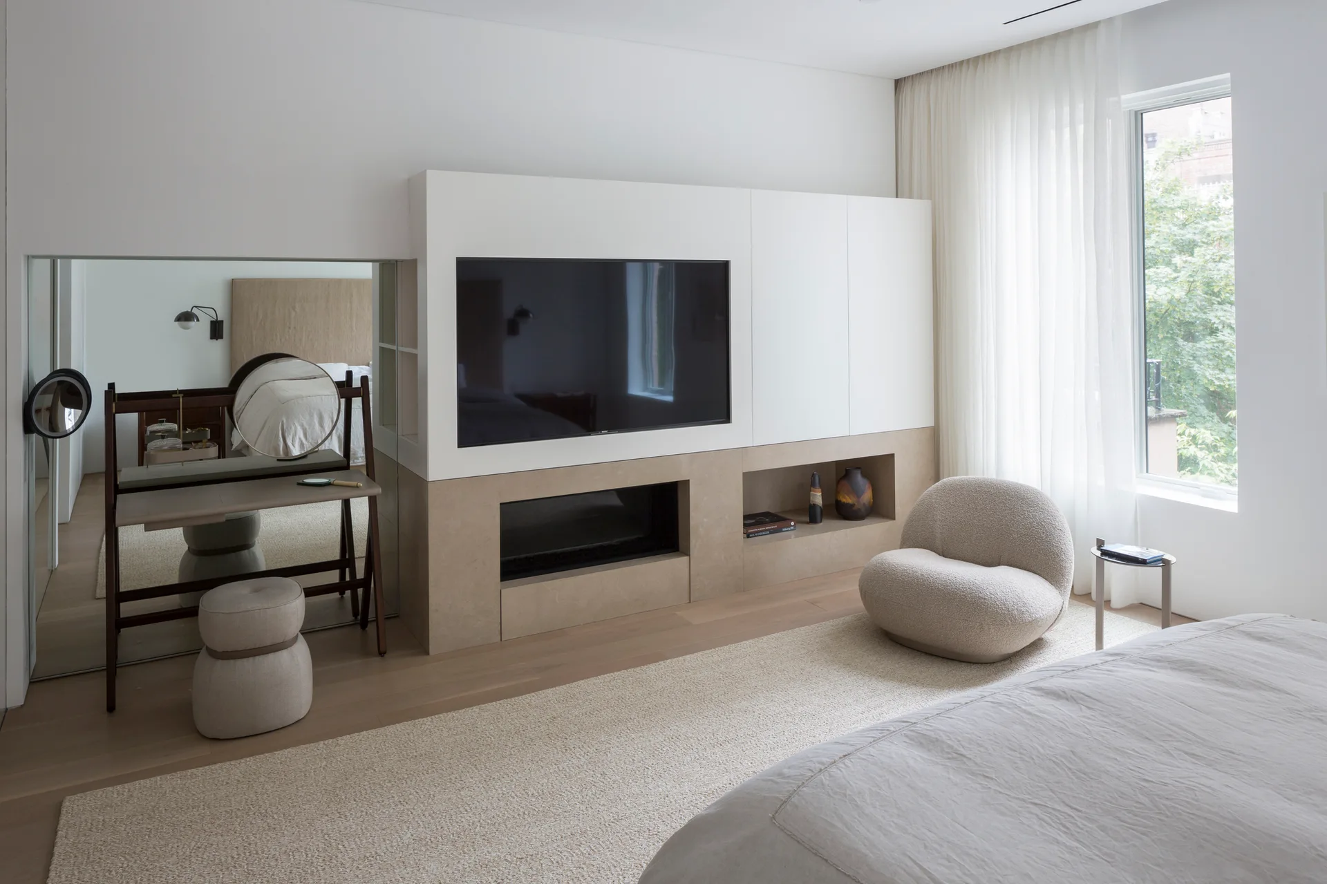 A modern bedroom features a custom white and beige media unit with an integrated fireplace, a round boucle armchair, and a vanity set against a mirrored wall.