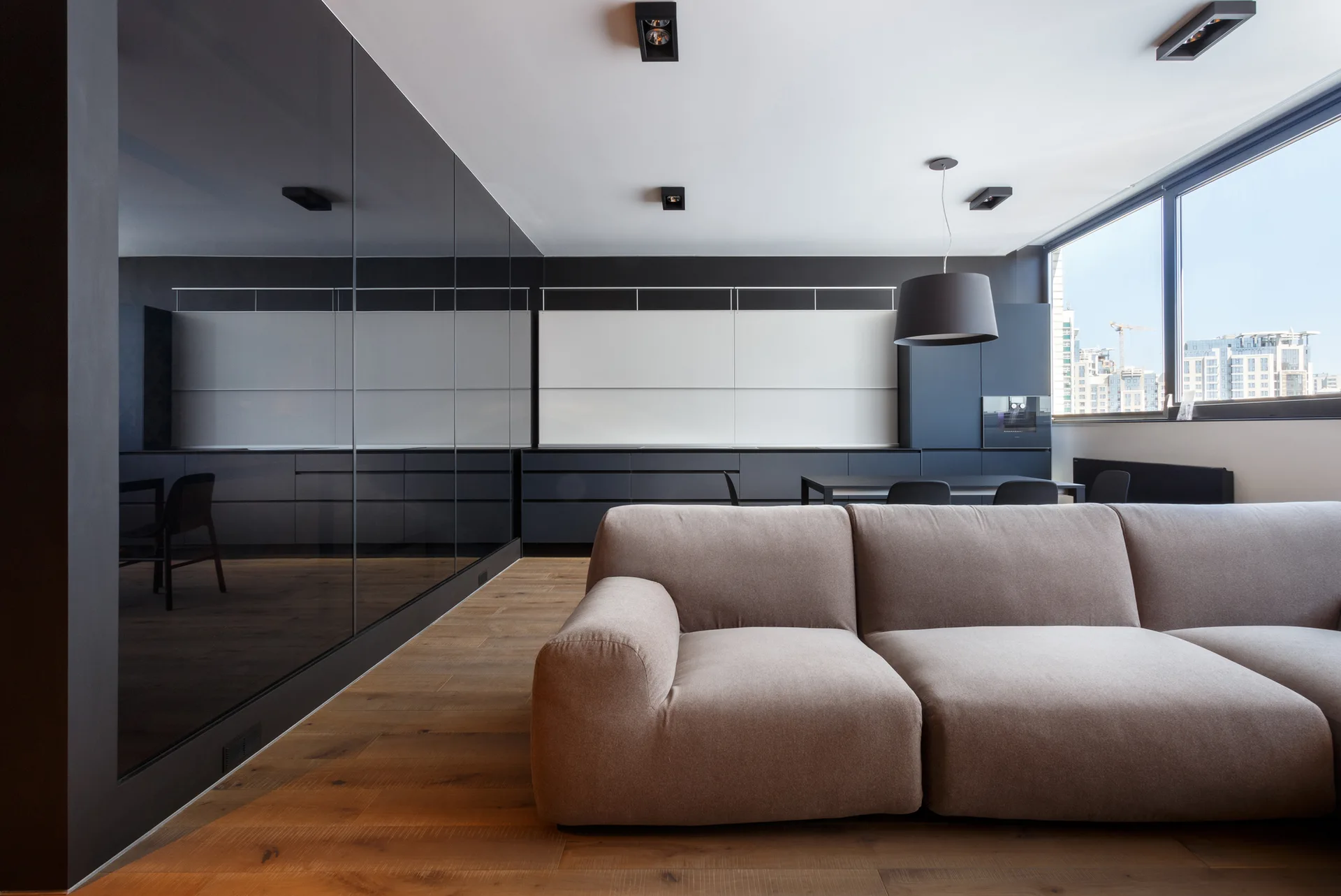 A modern open-plan living room features a low-profile tan sofa, medium wood floors, a dark mirrored wall, and minimalist black kitchen cabinets.
