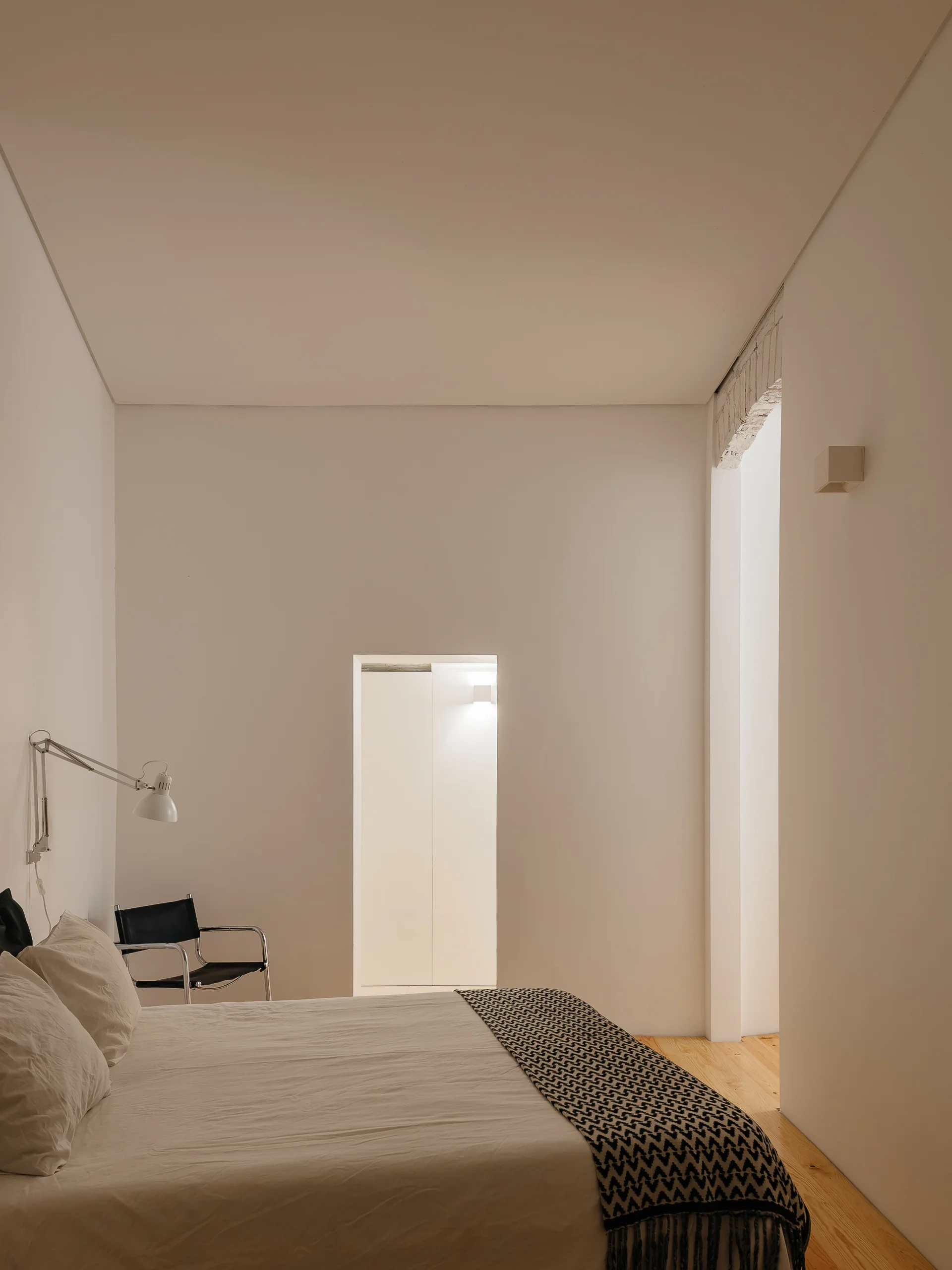 A minimalist white bedroom featuring a bed with white linens, a patterned throw, and a black leather armchair against plain white walls.
