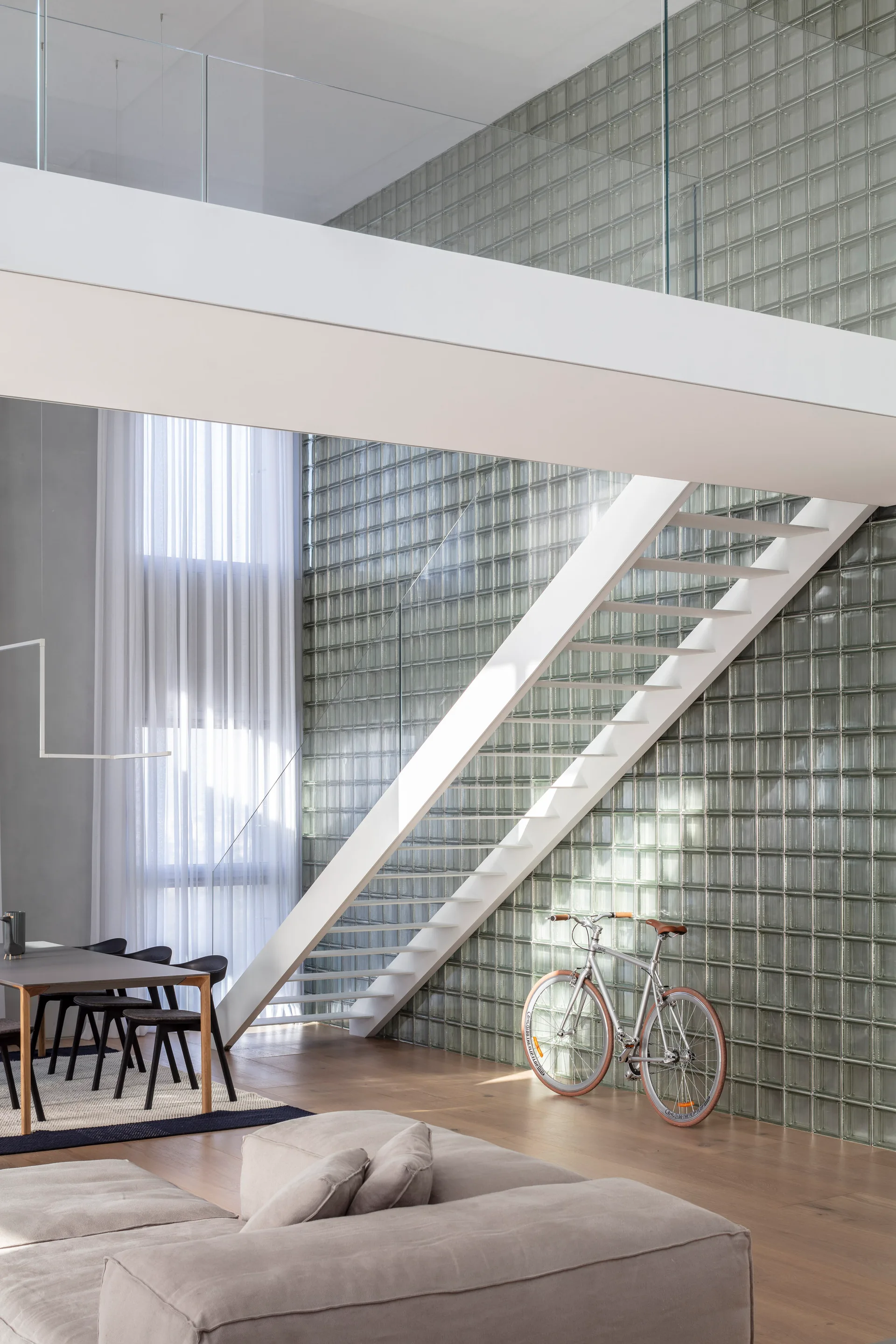 A white metal staircase ascends alongside a large glass block wall in a modern living room with wood flooring, a grey sofa, and a silver bicycle.