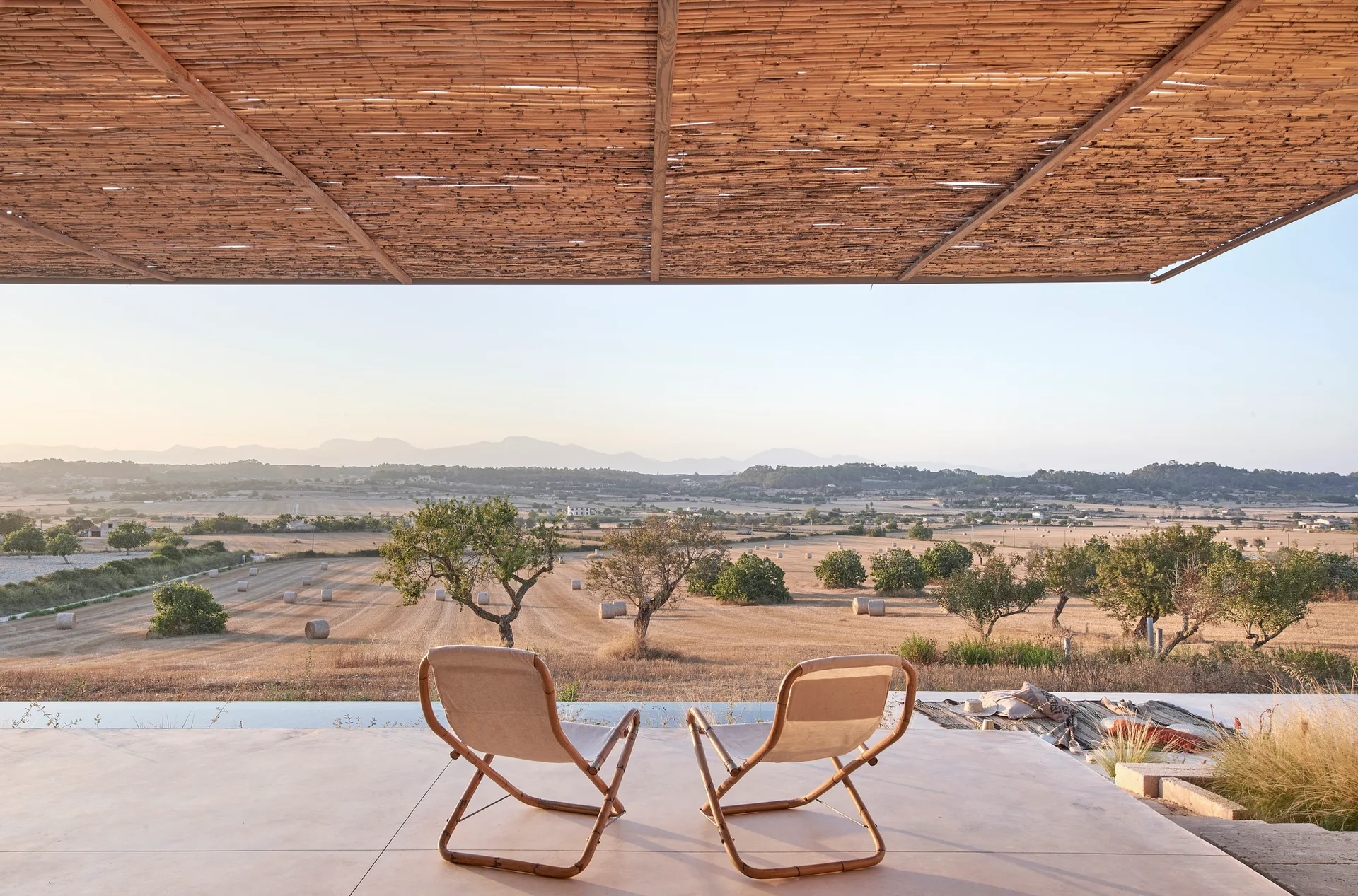 Two canvas sling chairs on a concrete patio shaded by a rustic bamboo pergola overlook a scenic field of hay bales.