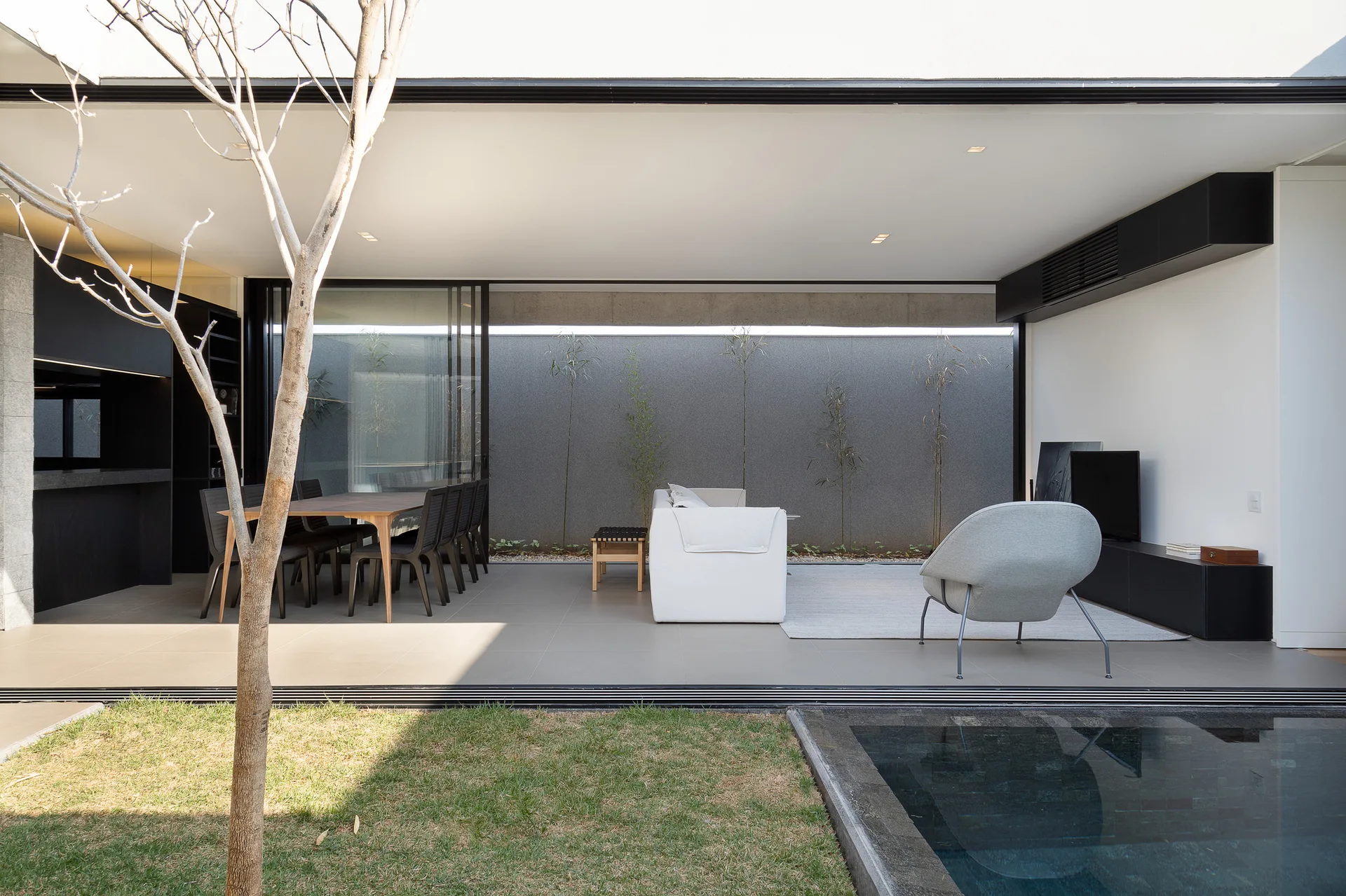 A minimalist patio features living and dining furniture, a large sliding glass wall, a grey tiled floor, and a swimming pool in the foreground.